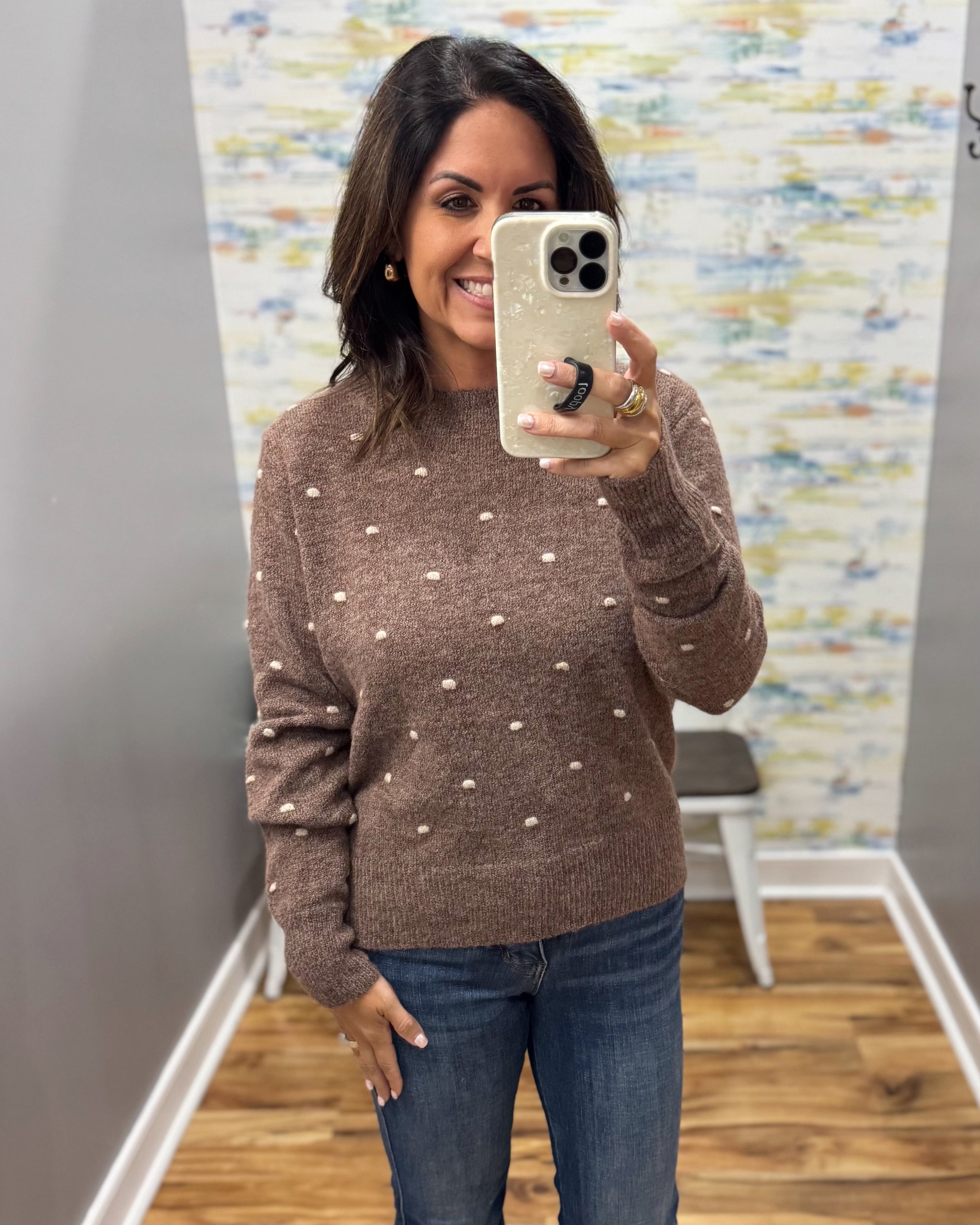 A woman takes a mirror selfie wearing a brown knit sweater with small white polka dots and blue jeans in a fitting room with wood floors and a patterned wall.