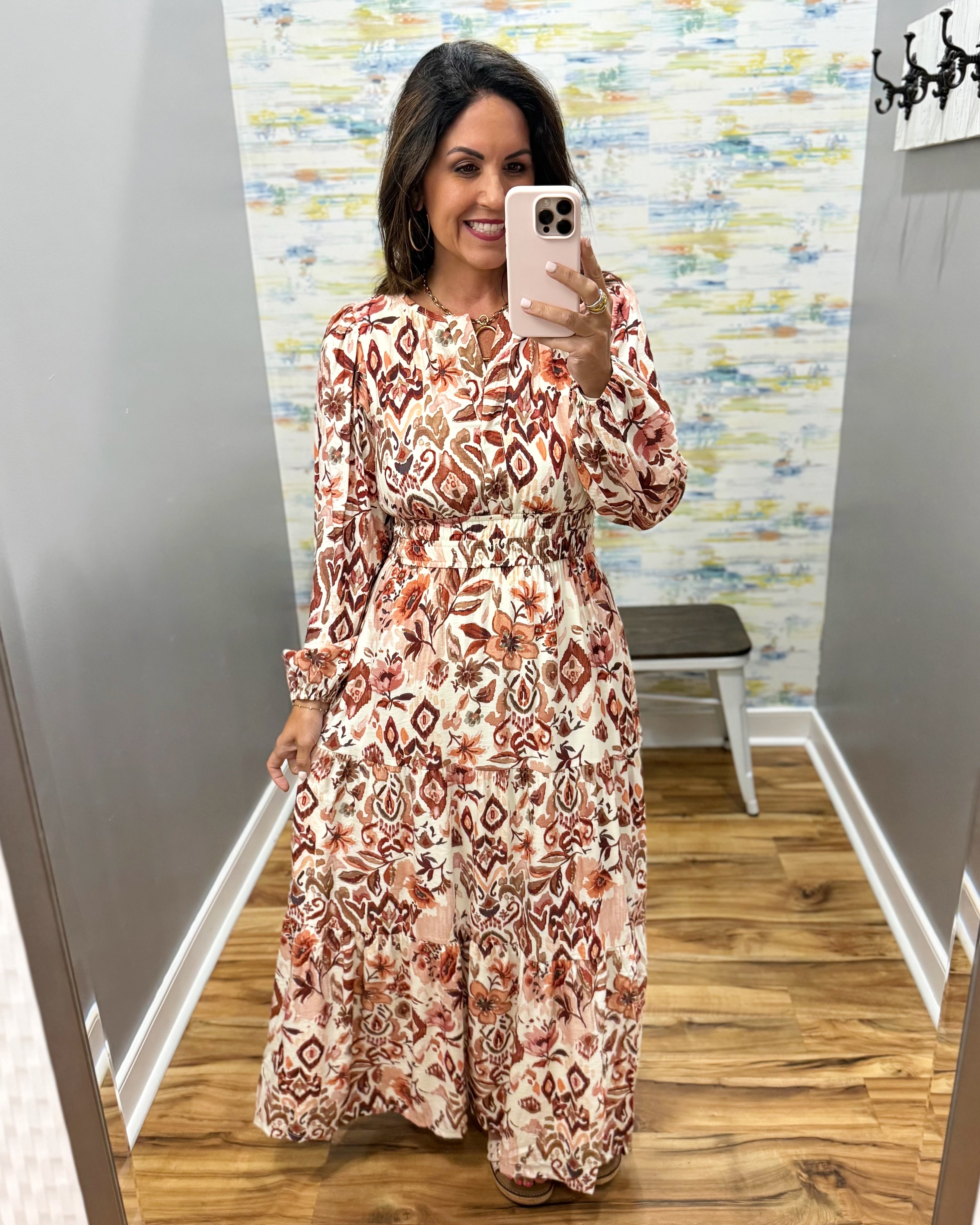 A woman takes a mirror selfie in a fitting room wearing a long-sleeve maxi dress with a brown, rust, and beige geometric ikat pattern on a cream background.