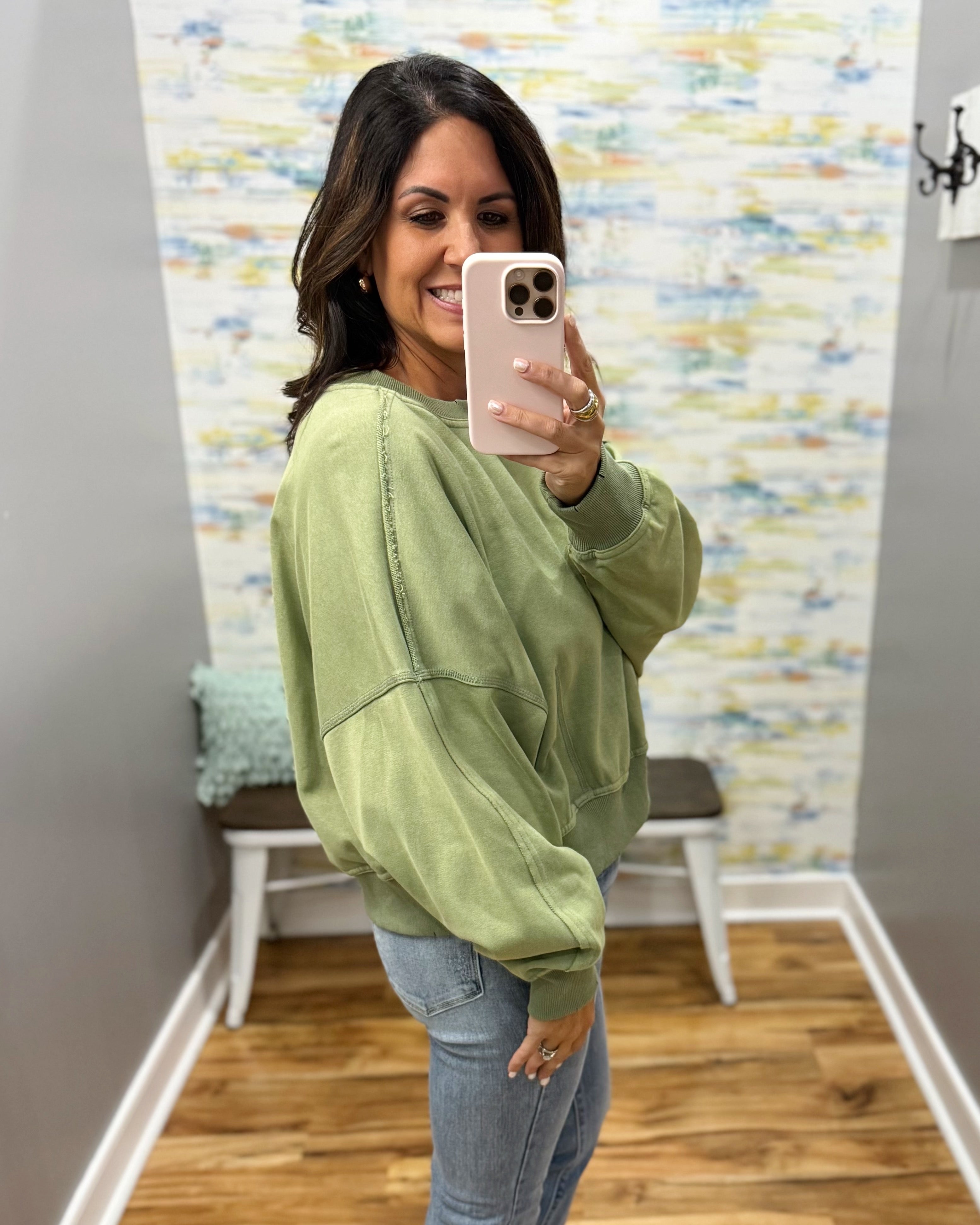 A woman takes a mirror selfie wearing an oversized sage green sweatshirt and light blue jeans in a home setting with a patterned wall and wood floor.