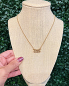 A gold necklace with a small floral or leafy pendant displayed on a beige fabric jewelry bust, with a hand holding the bust at the side against a green leafy background.