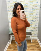 A woman takes a mirror selfie wearing a ribbed long-sleeve knit sweater in a warm rust orange color, paired with light-wash blue jeans. She is standing in a fitting room with a patterned wallpaper background.