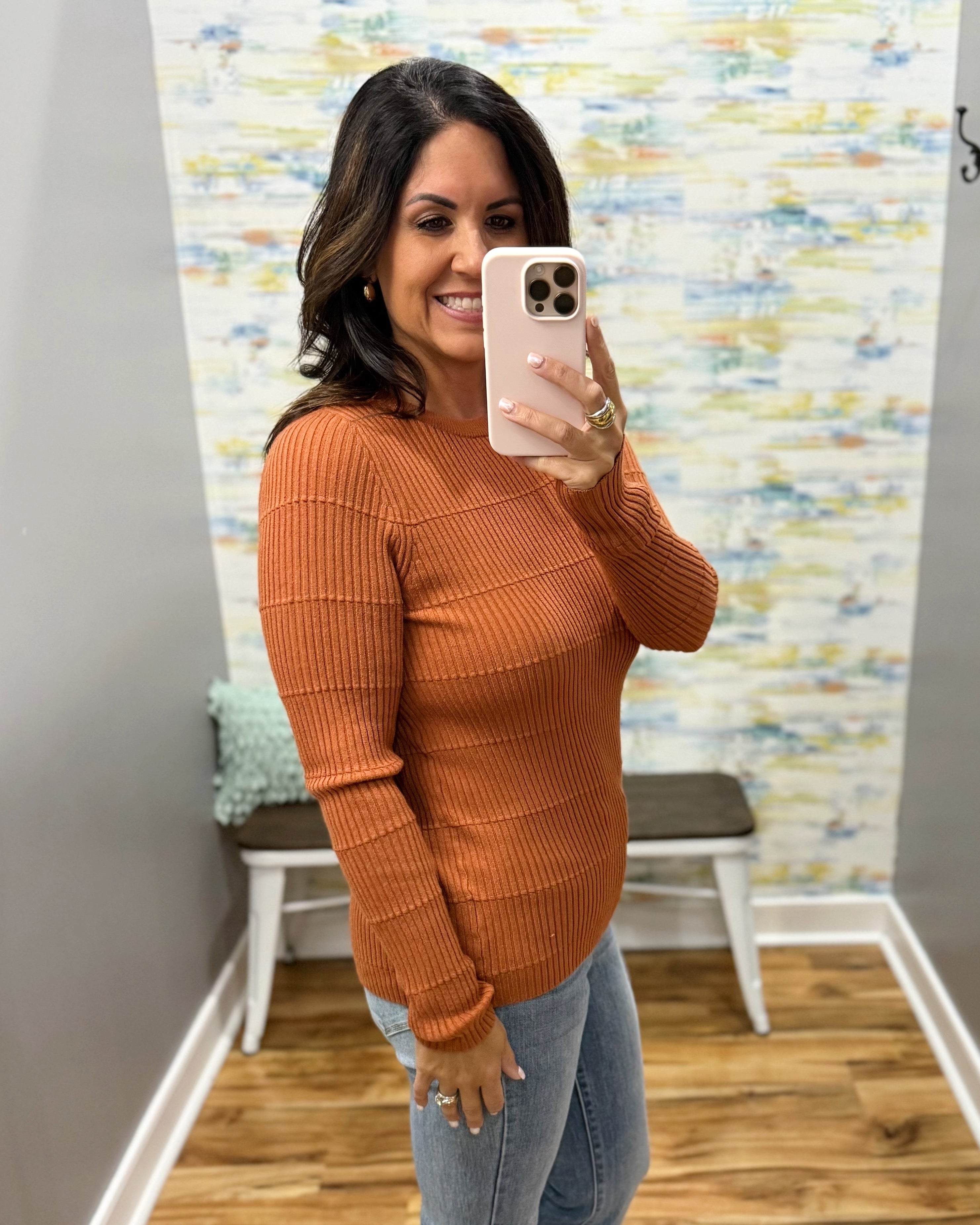 A woman takes a mirror selfie wearing a ribbed long-sleeve knit sweater in a warm rust orange color, paired with light-wash blue jeans. She is standing in a fitting room with a patterned wallpaper background.