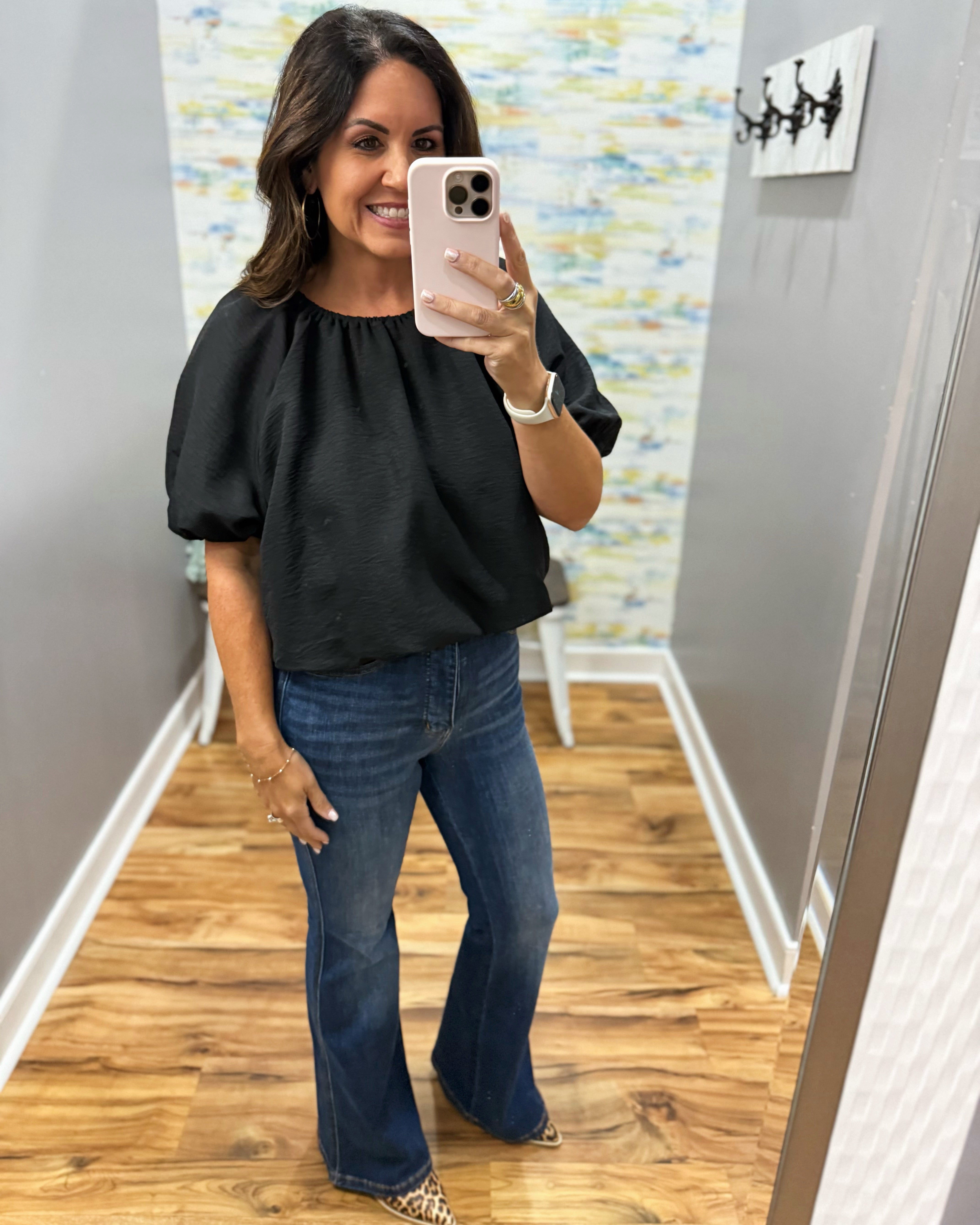 A woman takes a mirror selfie in a fitting room or hallway, wearing a black puff-sleeve top and blue flare jeans. The background features gray walls, a wood floor, and abstract wall art.