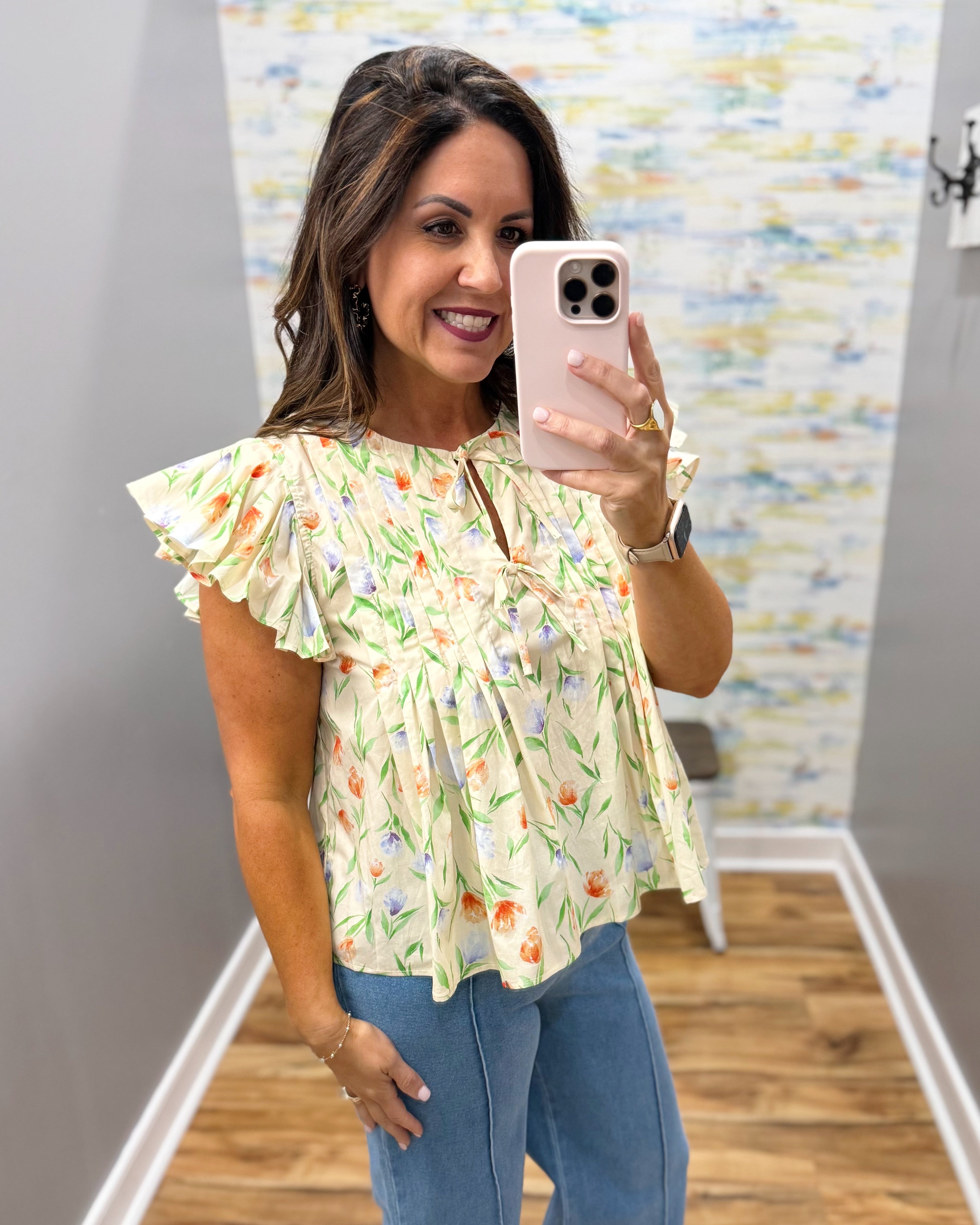 Woman taking a mirror selfie wearing a floral top and blue jeans.