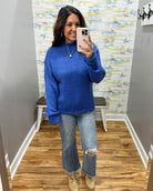 A woman takes a mirror selfie wearing a vibrant royal blue ribbed knit sweater, light-wash distressed jeans, and beige lace-up boots in a fitting room with wood floors and a patterned wallpaper background.