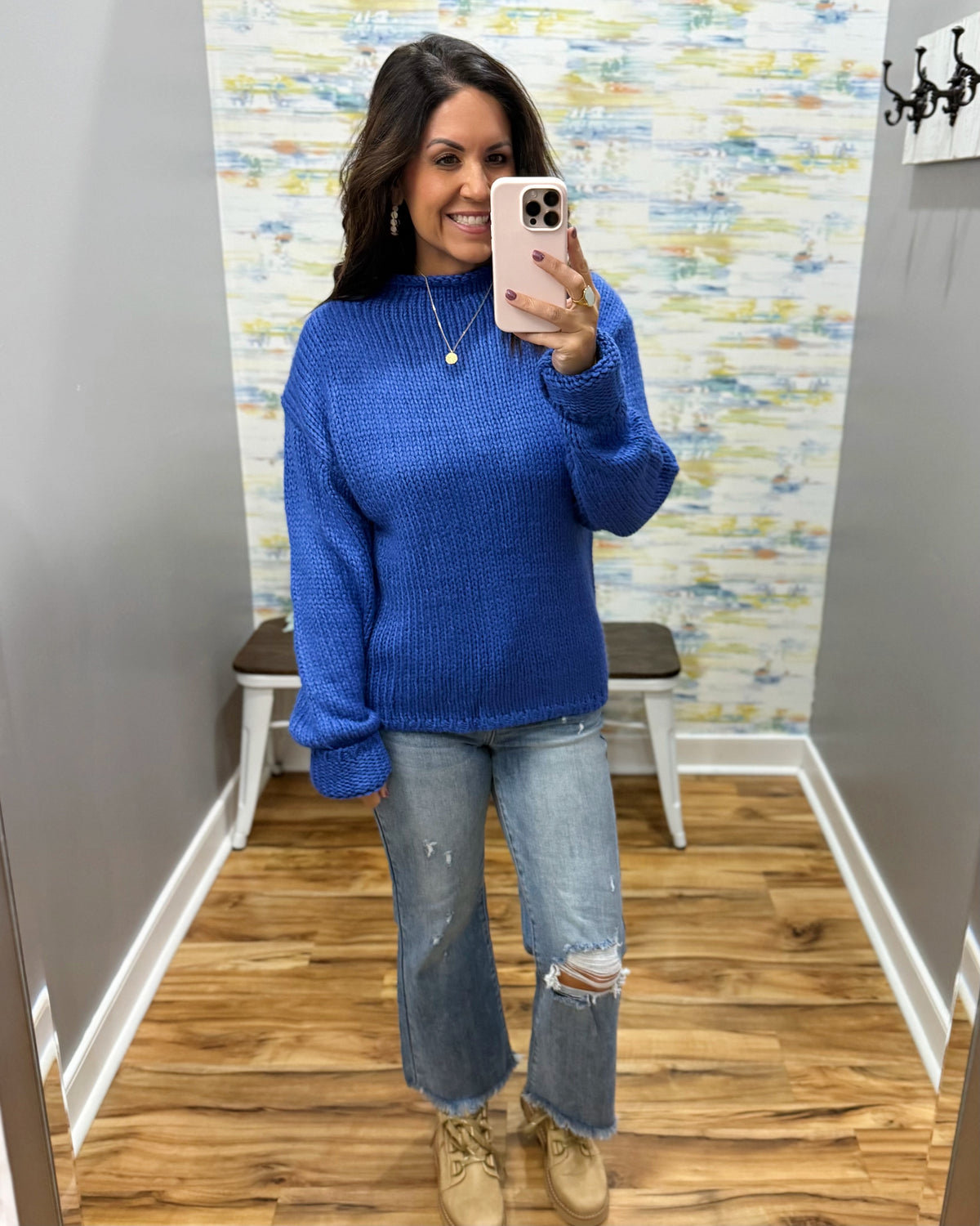 A woman takes a mirror selfie wearing a vibrant royal blue ribbed knit sweater, light-wash distressed jeans, and beige lace-up boots in a fitting room with wood floors and a patterned wallpaper background.