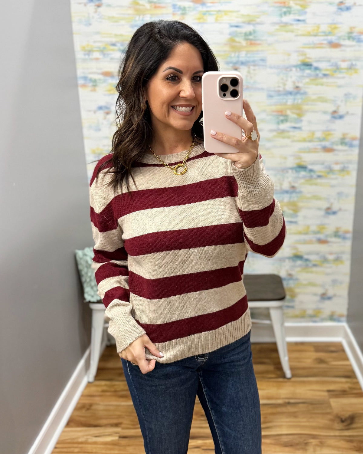 A woman takes a mirror selfie wearing a cream and burgundy striped knit sweater, blue jeans, and a gold necklace in an indoor room with a patterned wall and wood floor.