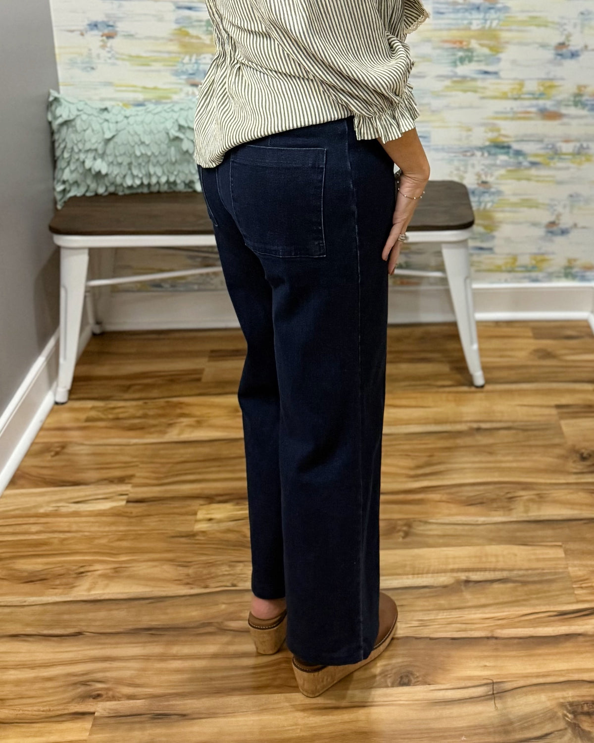 A person shown from the back wearing dark navy wide-leg pants, a cream and green striped top, and tan wedge sandals in an indoor room with wood floors and a bench.