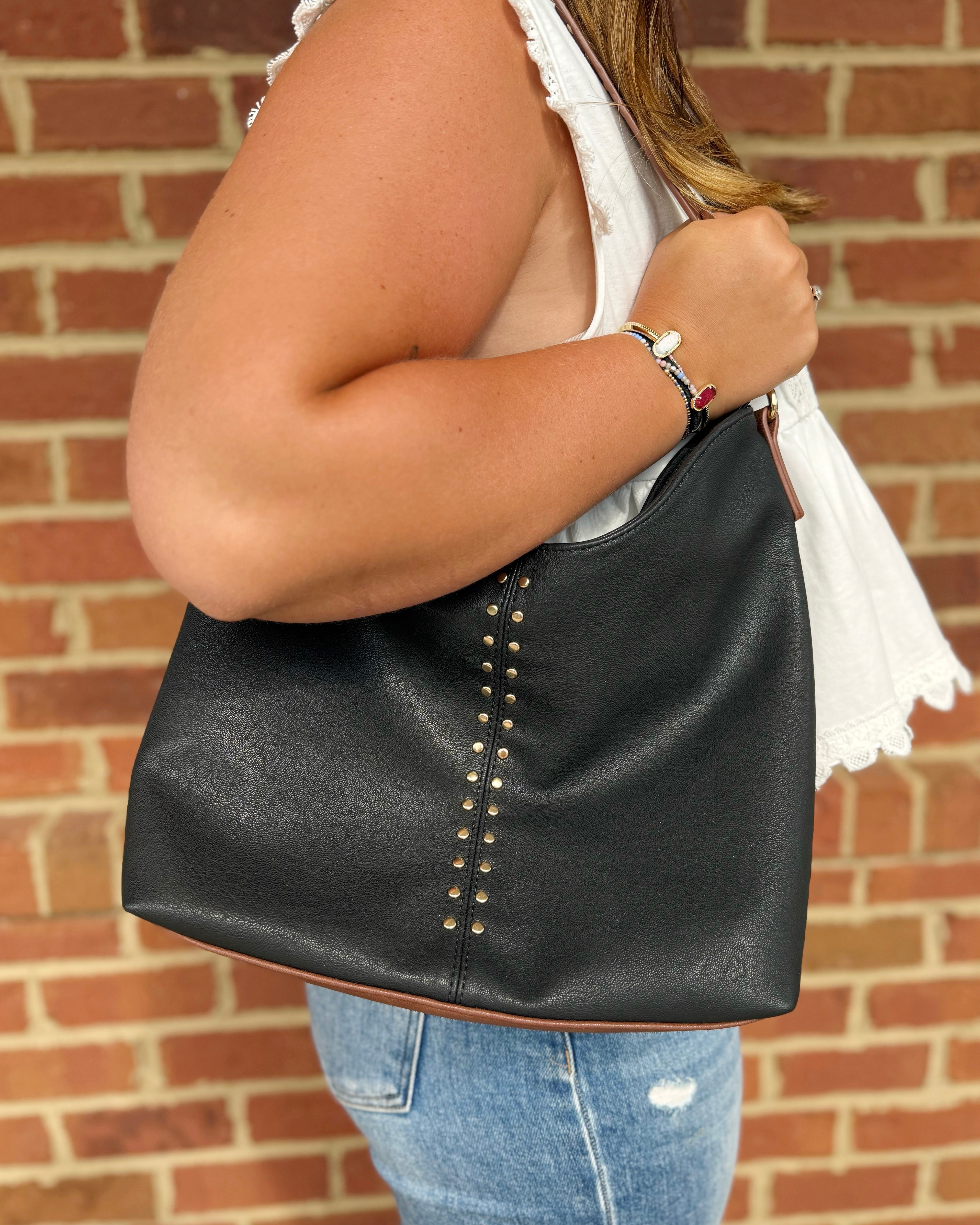 A woman wearing a white sleeveless top and blue jeans carries a large black faux-leather hobo shoulder bag with gold-tone stud detailing, set against a red brick wall background.