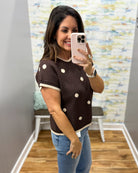 A woman takes a mirror selfie in a fitting room wearing a brown short-sleeve knit sweater with white pom-pom dots and cream trim, paired with blue jeans.