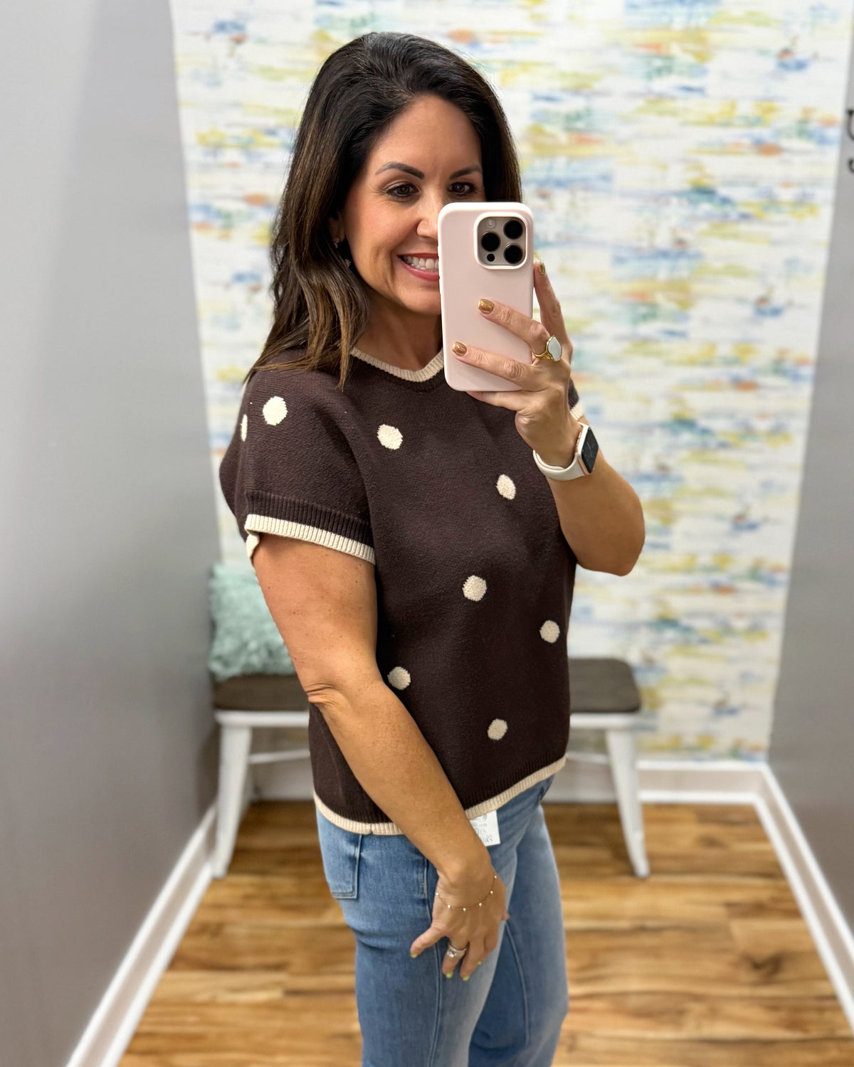 A woman takes a mirror selfie in a fitting room wearing a brown short-sleeve knit sweater with white pom-pom dots and cream trim, paired with blue jeans.