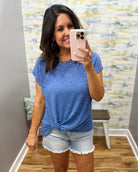 Woman taking a mirror selfie wearing a blue t-shirt and denim shorts in a room with a patterned wall.
