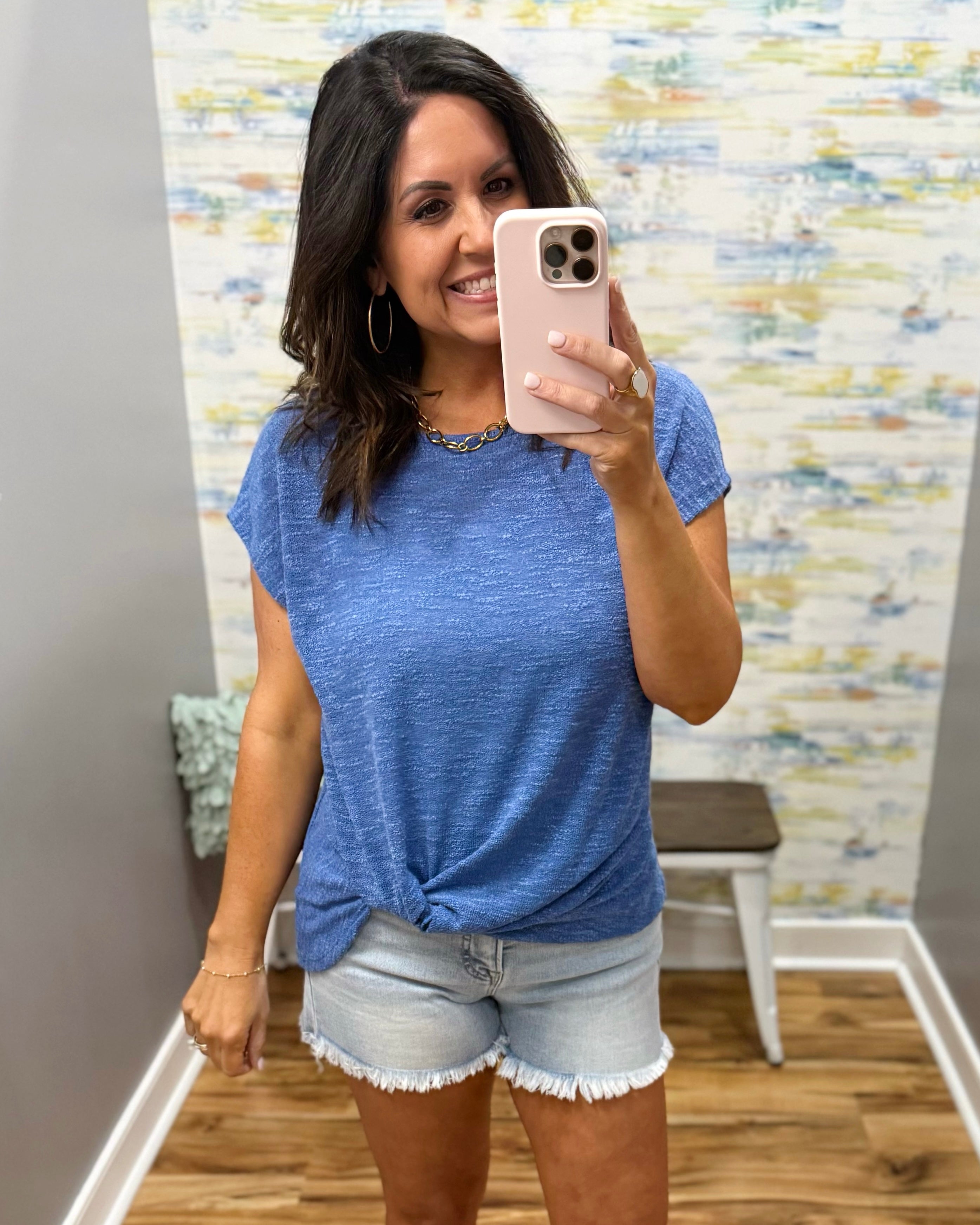 Woman taking a mirror selfie wearing a blue t-shirt and denim shorts in a room with a patterned wall.