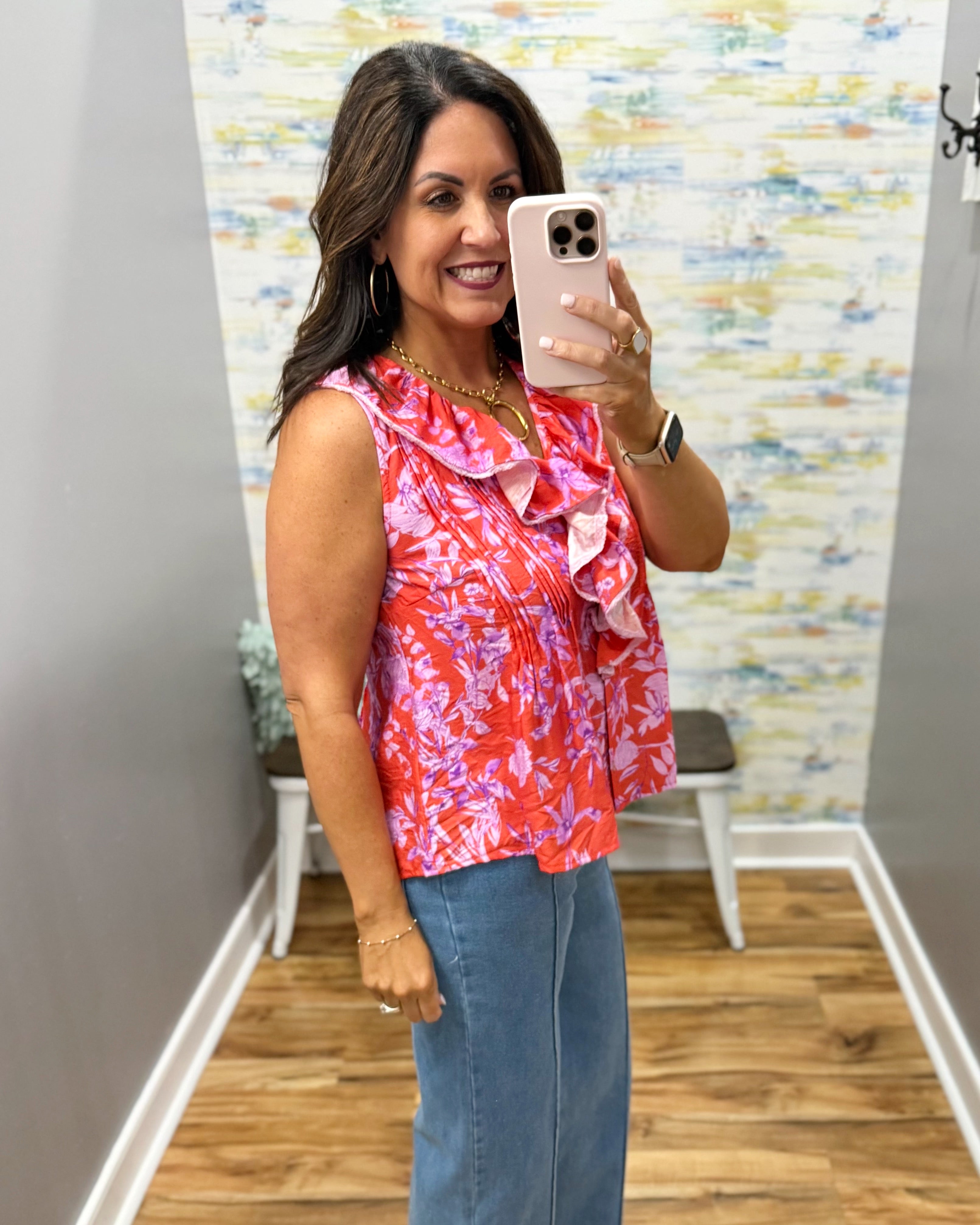 Woman taking a mirror selfie wearing a colorful sleeveless top and jeans in a room with a patterned wall.