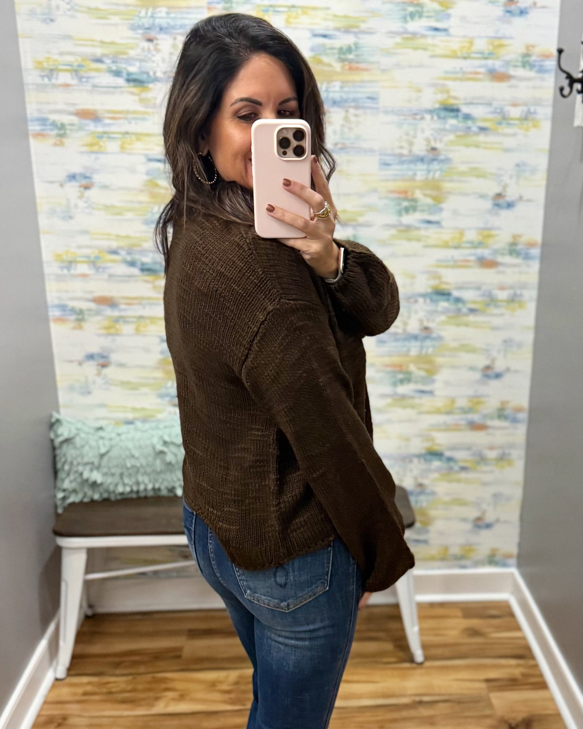 A woman takes a mirror selfie wearing a brown textured knit sweater and blue jeans in front of a colorful abstract wallpapered wall.