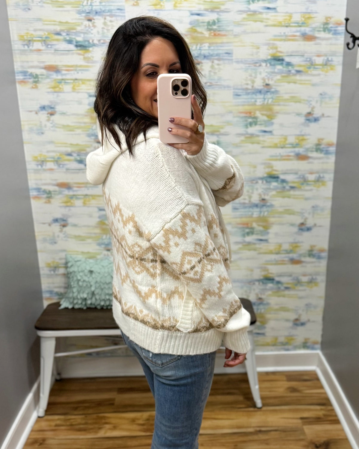 A woman takes a mirror selfie wearing a cream and tan patterned knit sweater with a hood, paired with blue jeans, in front of a patterned wallpapered wall.