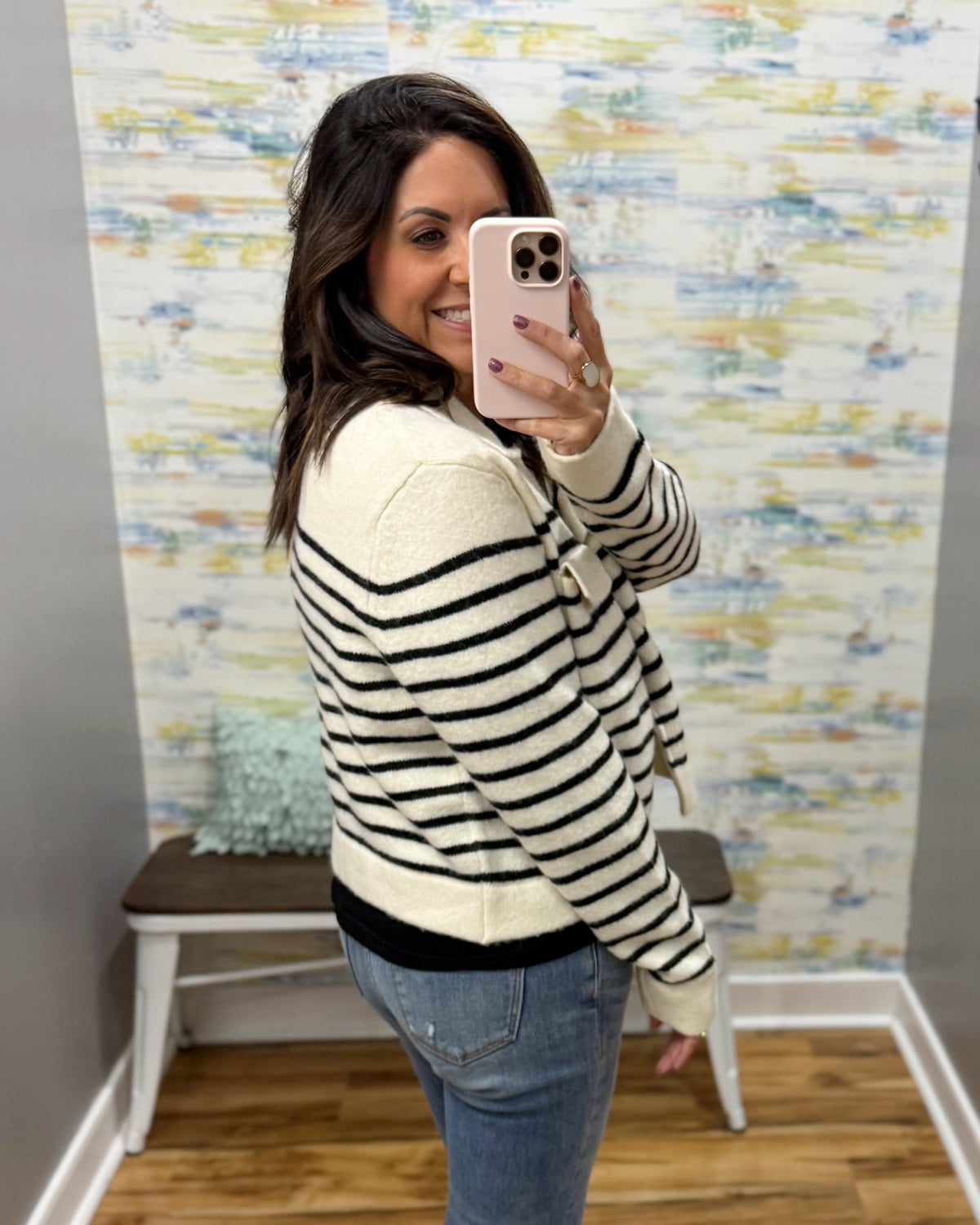 A woman takes a mirror selfie wearing a cream and black horizontally striped knit sweater, blue jeans, and a light pink phone case in front of a patterned wallpapered wall.
