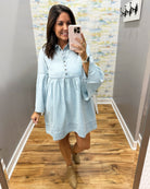 A woman takes a mirror selfie wearing a light blue long-sleeve button-front dress with a gathered waist and flared bell sleeves, paired with tan ankle boots in a home hallway.