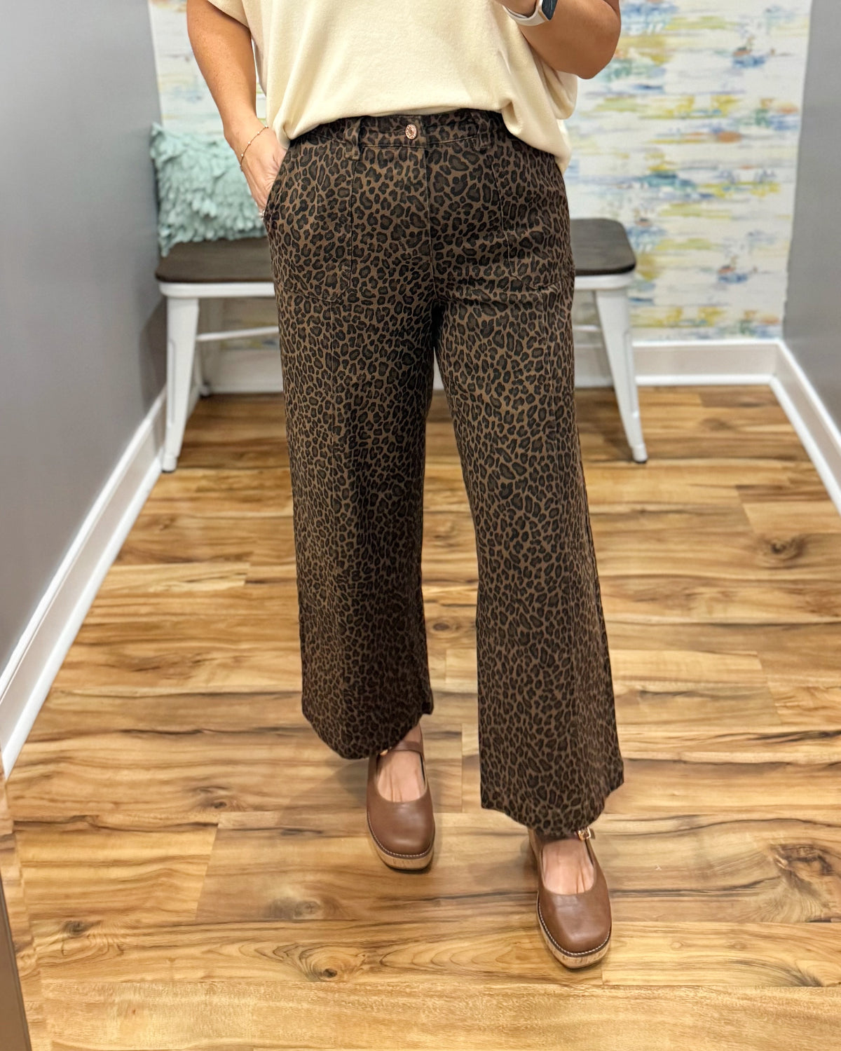 A person wearing brown leopard-print wide-leg pants, a cream top, and brown shoes, standing in a room with a wood floor and a bench in the background.