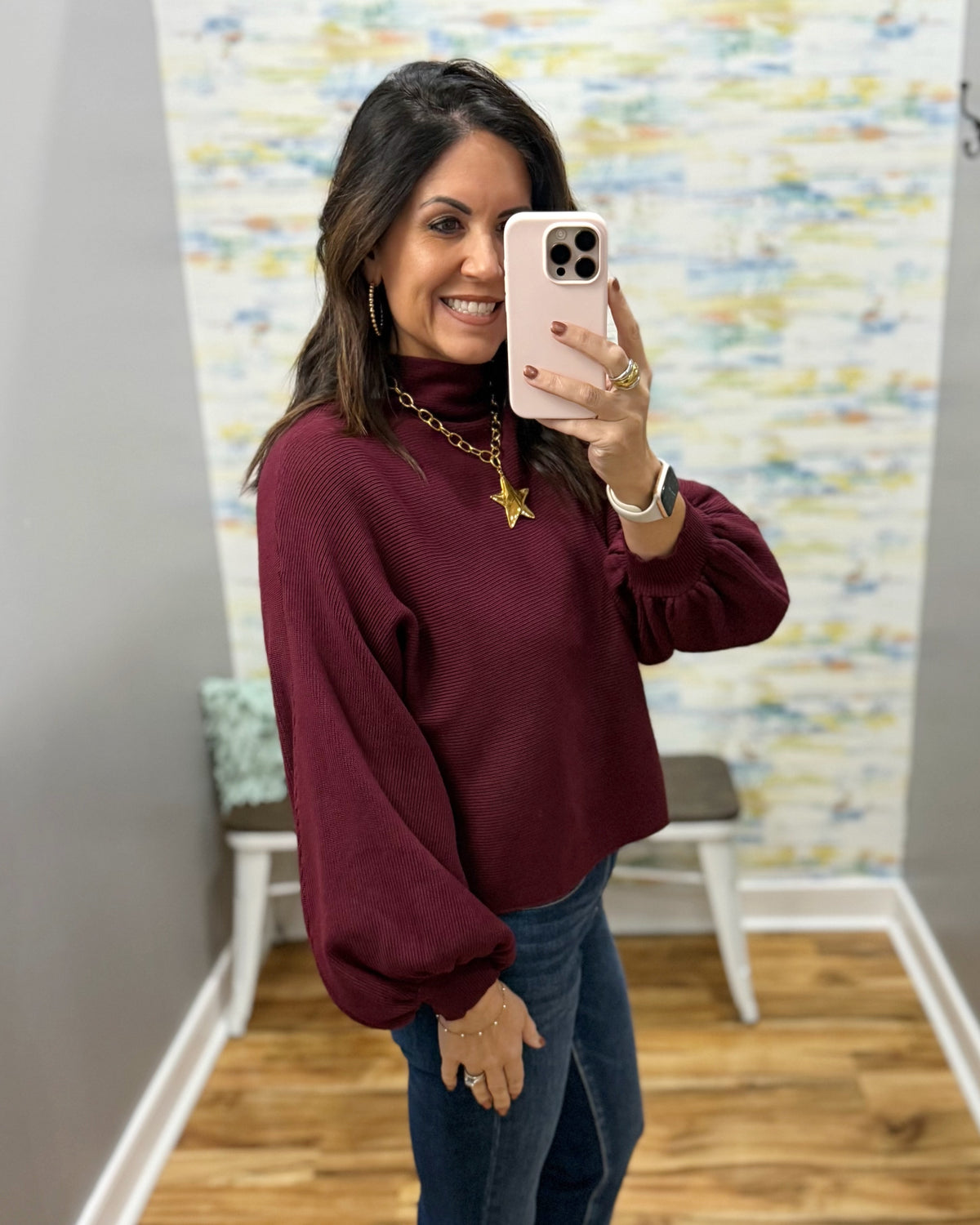 A woman takes a mirror selfie wearing a maroon long-sleeve turtleneck sweater and blue jeans in a fitting room with a colorful abstract wall mural.