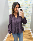 A woman takes a mirror selfie wearing a dark purple long-sleeve blouse with a small white polka dot pattern and blue jeans.