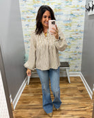 A woman takes a mirror selfie in a fitting room wearing a beige and white vertically striped long-sleeve blouse with ruffles, blue flare jeans, and tan pointed-toe heels.