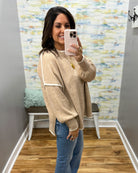 A woman takes a mirror selfie in a fitting room wearing a beige knit sweater with white contrast trim and blue jeans.