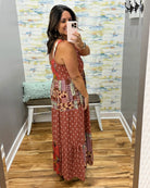 A woman takes a mirror selfie in a fitting room wearing a rust-colored maxi dress with a patchwork print and white geometric patterns. The fitting room has wood floors, a bench with a mint green pillow, and a blue and yellow abstract patterned wall.