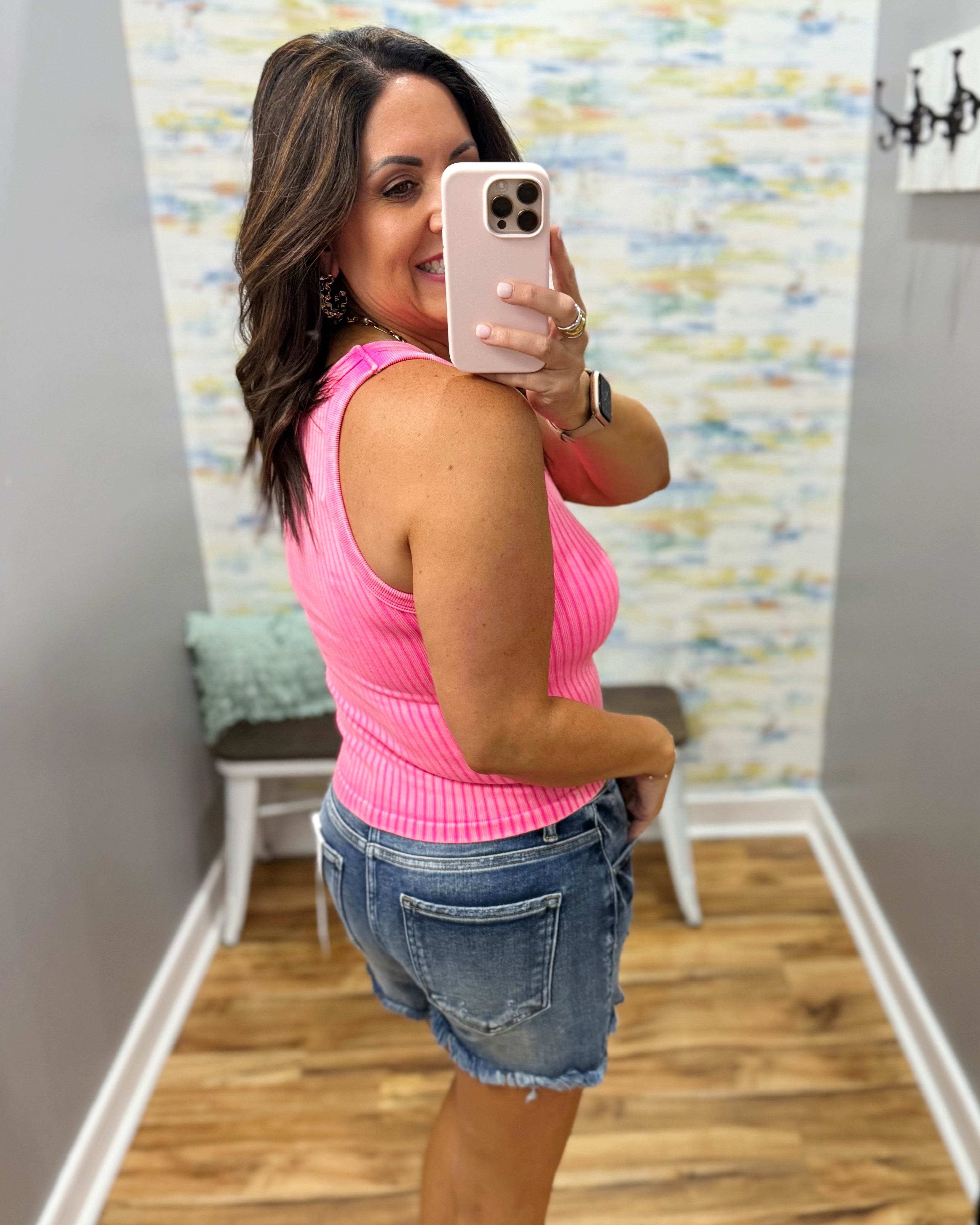 Woman taking a mirror selfie wearing a pink sleeveless top and denim shorts in a room with a colorful wall.