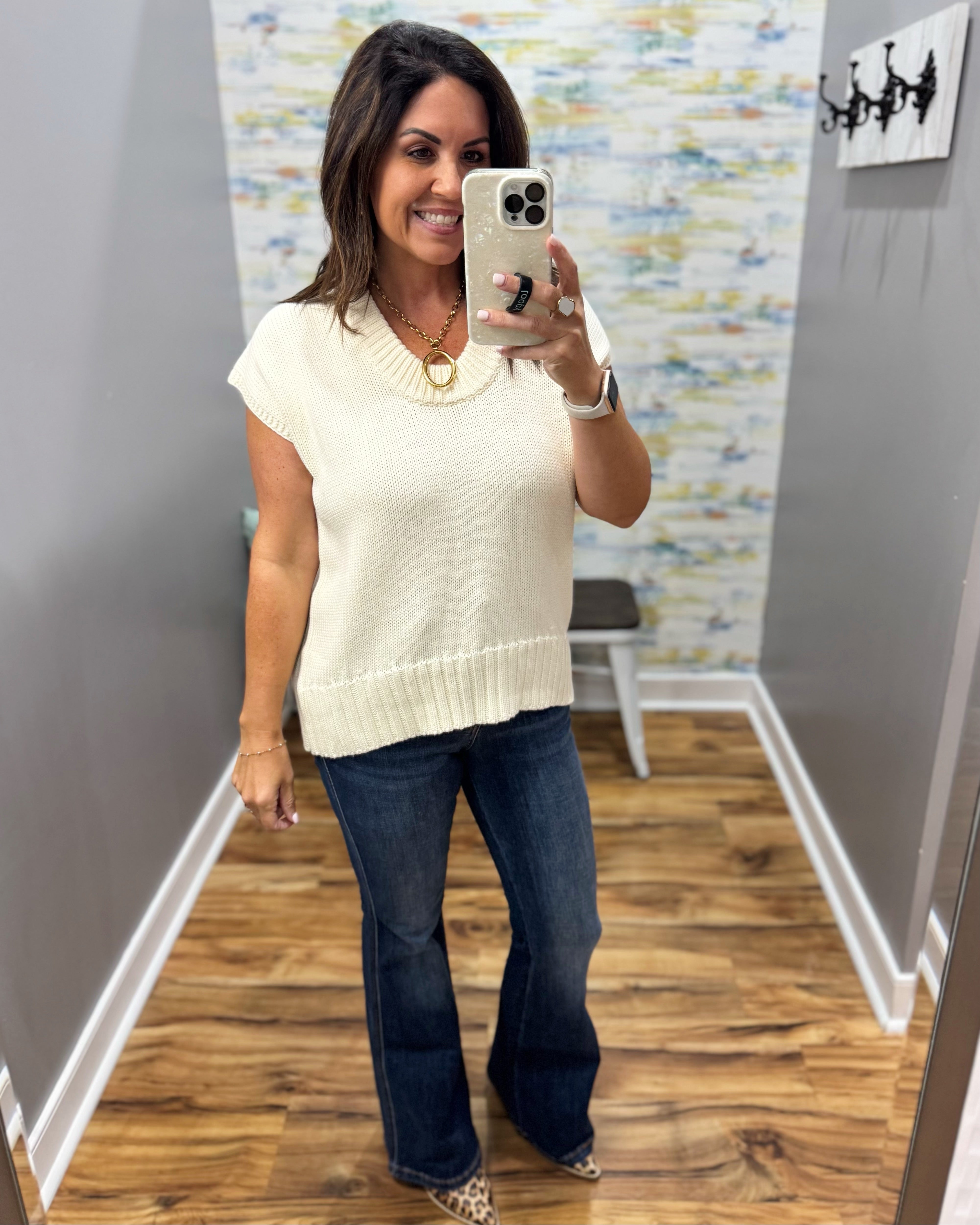 A woman takes a mirror selfie in a fitting room wearing a cream-colored sleeveless knit top and dark blue flare jeans.