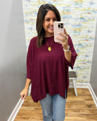 A woman takes a mirror selfie wearing a loose-fitting, three-quarter sleeve top in deep burgundy with light blue jeans. The background shows a gray wall, a patterned wallpaper, and a wood floor.