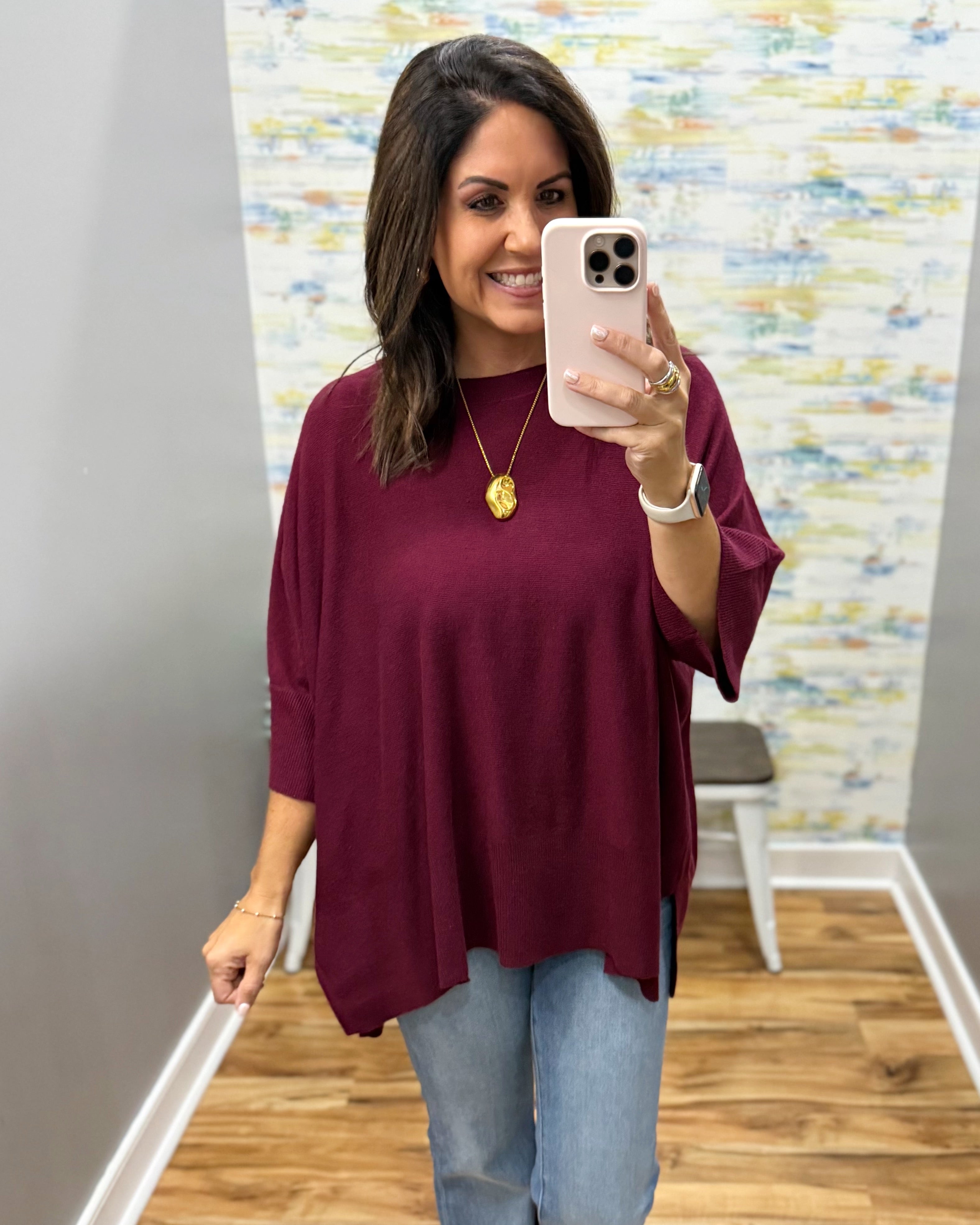 A woman takes a mirror selfie wearing a loose-fitting, three-quarter sleeve top in deep burgundy with light blue jeans. The background shows a gray wall, a patterned wallpaper, and a wood floor.