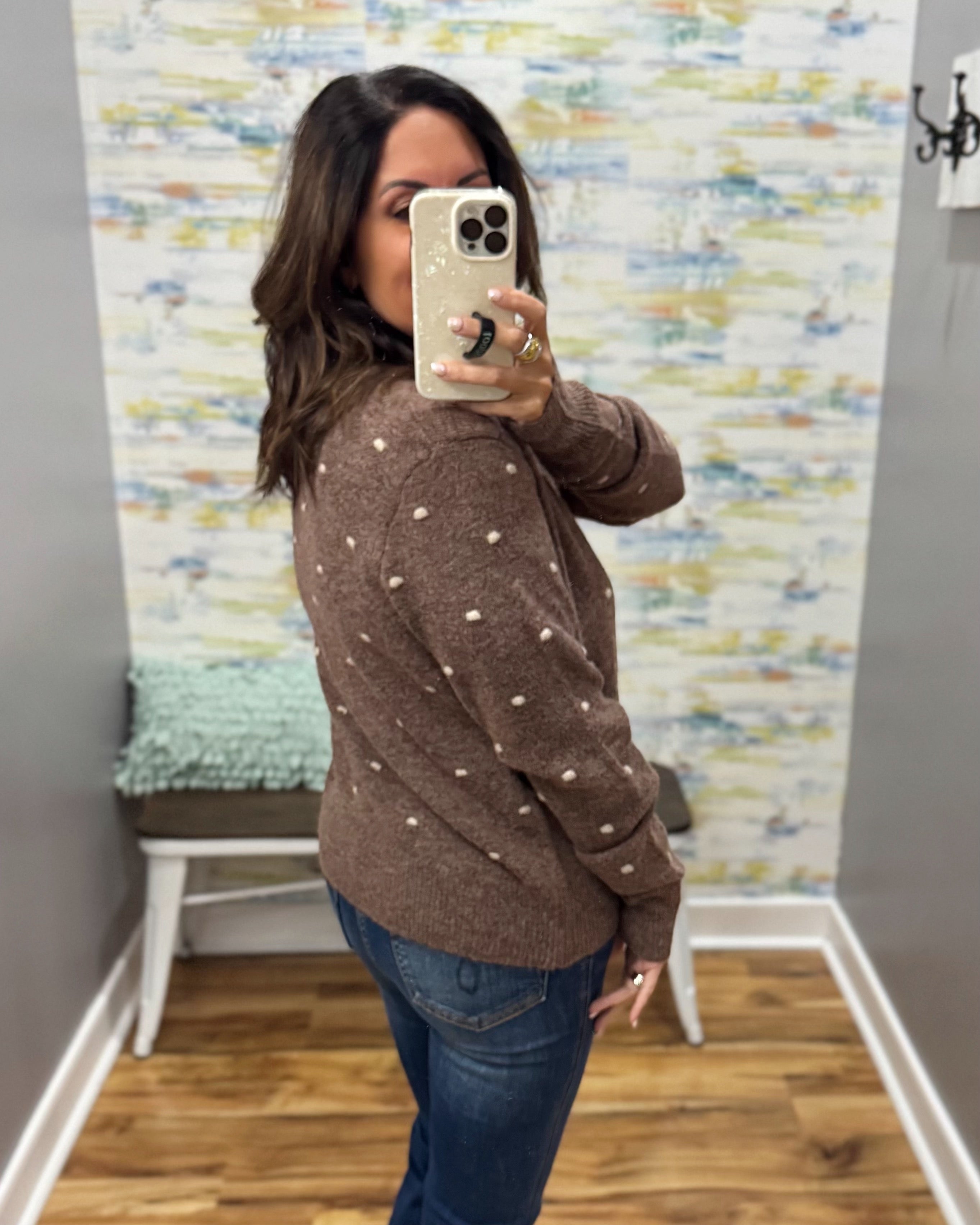 A woman takes a mirror selfie wearing a brown knit sweater with small white polka dots and blue jeans, standing in a room with a patterned wallpaper background.