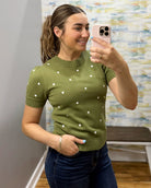 A woman takes a mirror selfie wearing a short-sleeve olive green knit sweater with white polka dots and dark blue jeans.