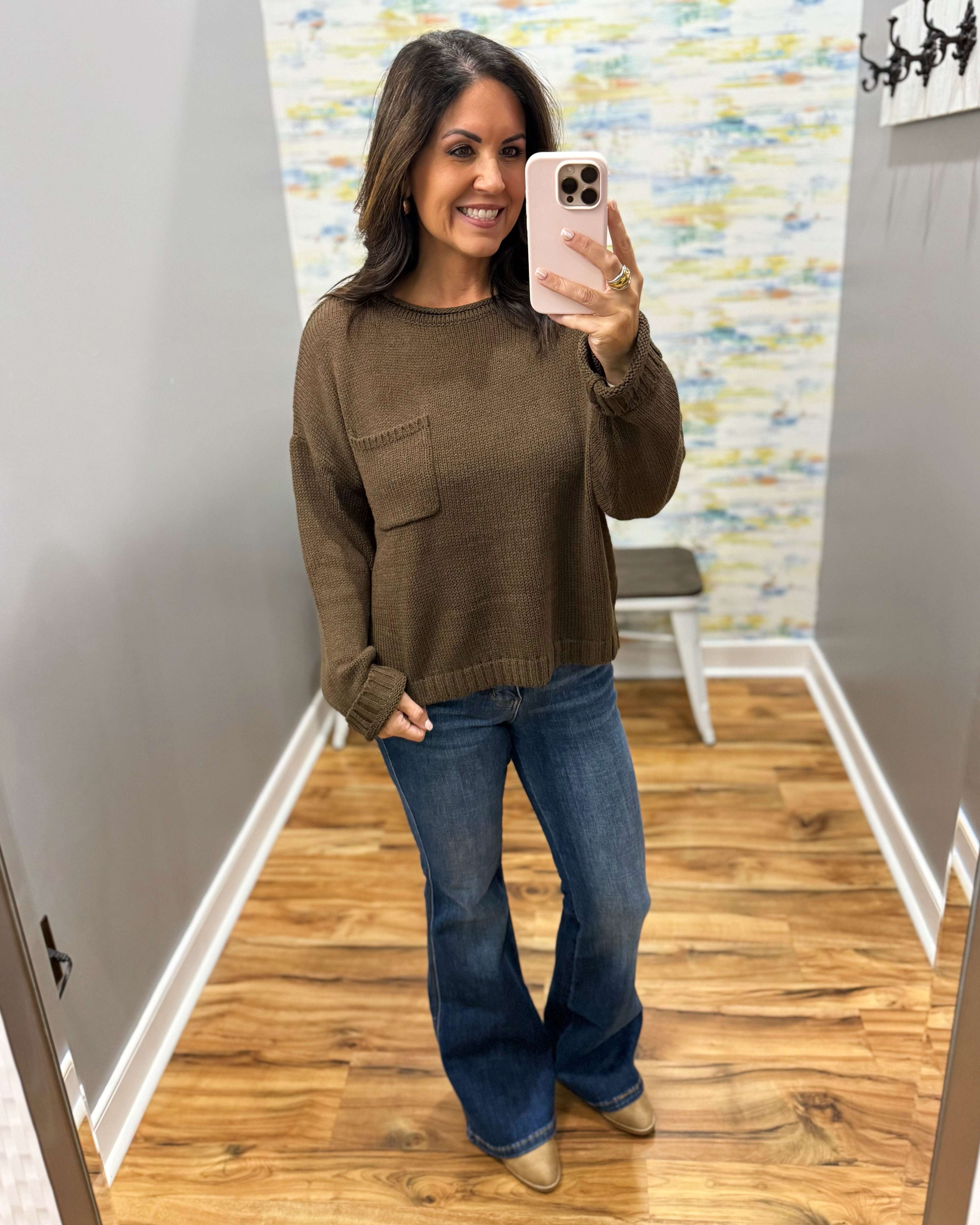 A woman takes a mirror selfie in a fitting room wearing a brown knit crewneck sweater with a chest pocket and blue flare jeans.