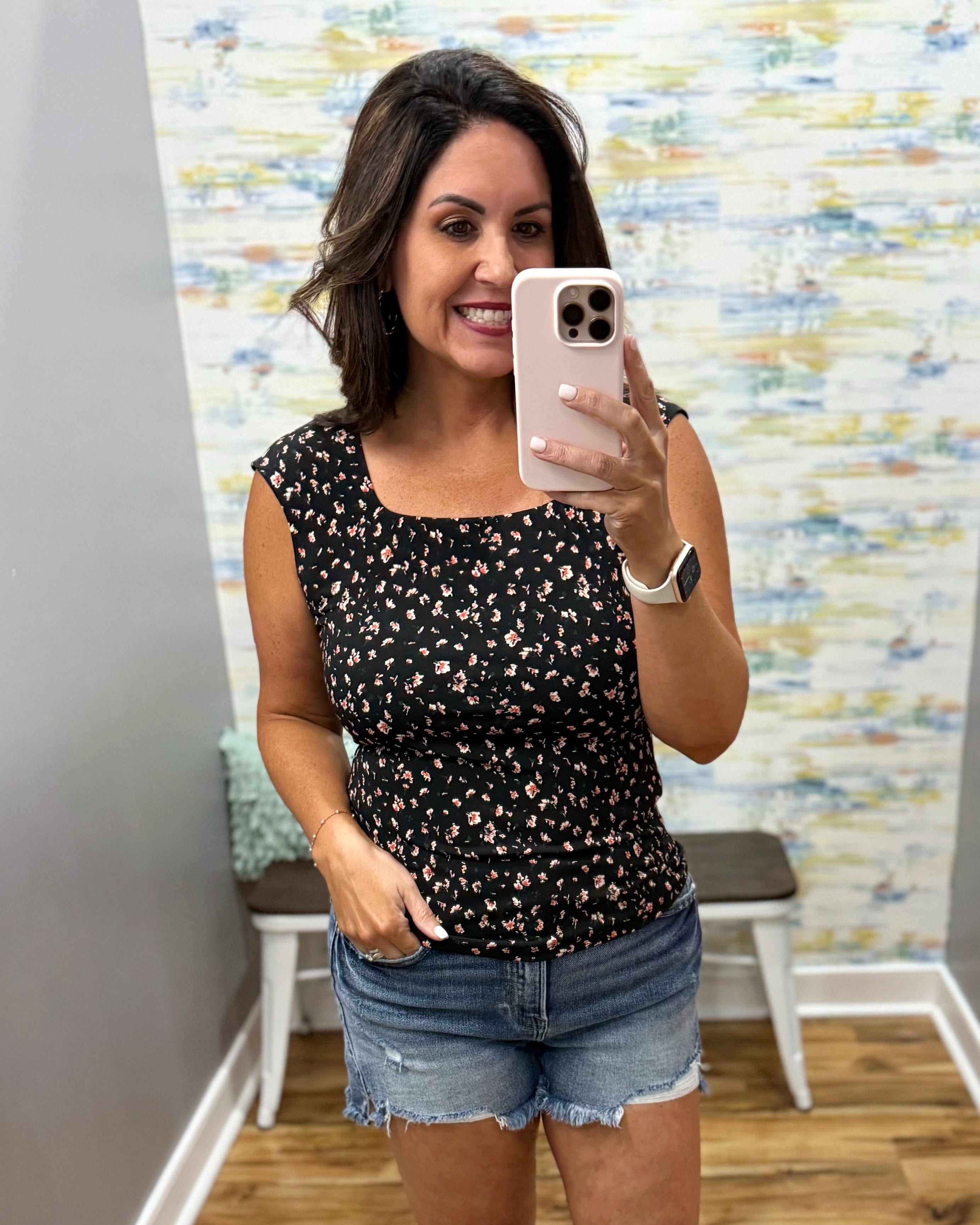 Woman taking a mirror selfie wearing a black floral top and denim shorts in a room with colorful wall art.