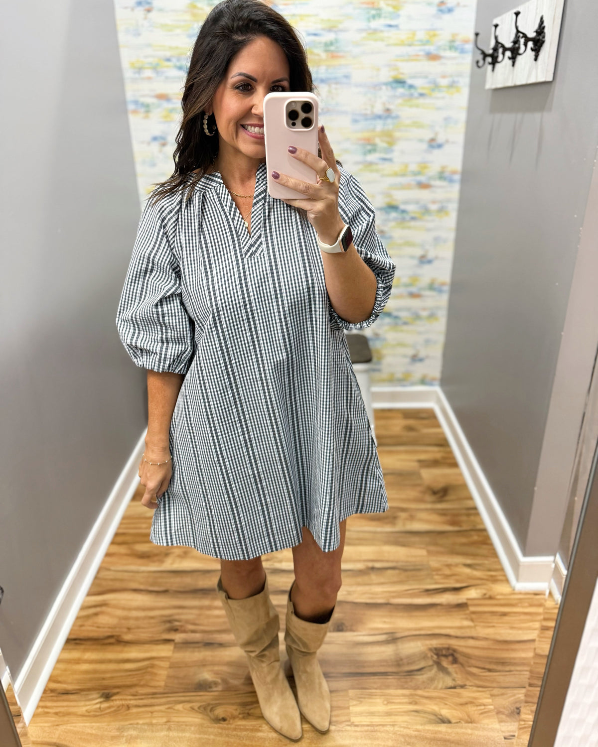 A woman takes a mirror selfie wearing a blue and white vertically striped mini shirt dress with puff sleeves and beige ankle boots in a fitting room.