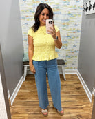 A woman takes a mirror selfie in a fitting room wearing a yellow short-sleeve peplum top and wide-leg blue jeans.