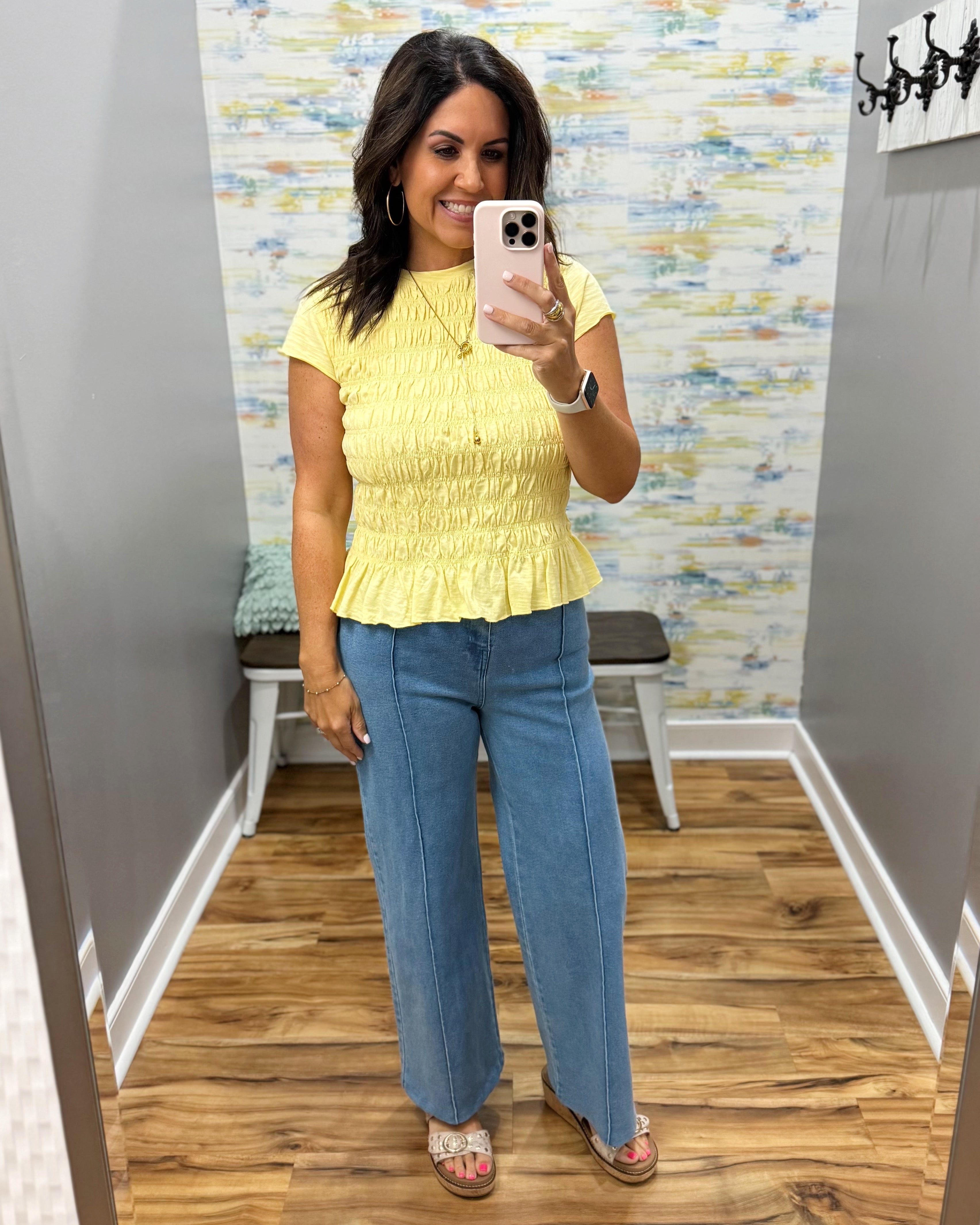 A woman takes a mirror selfie in a fitting room wearing a yellow short-sleeve peplum top and wide-leg blue jeans.