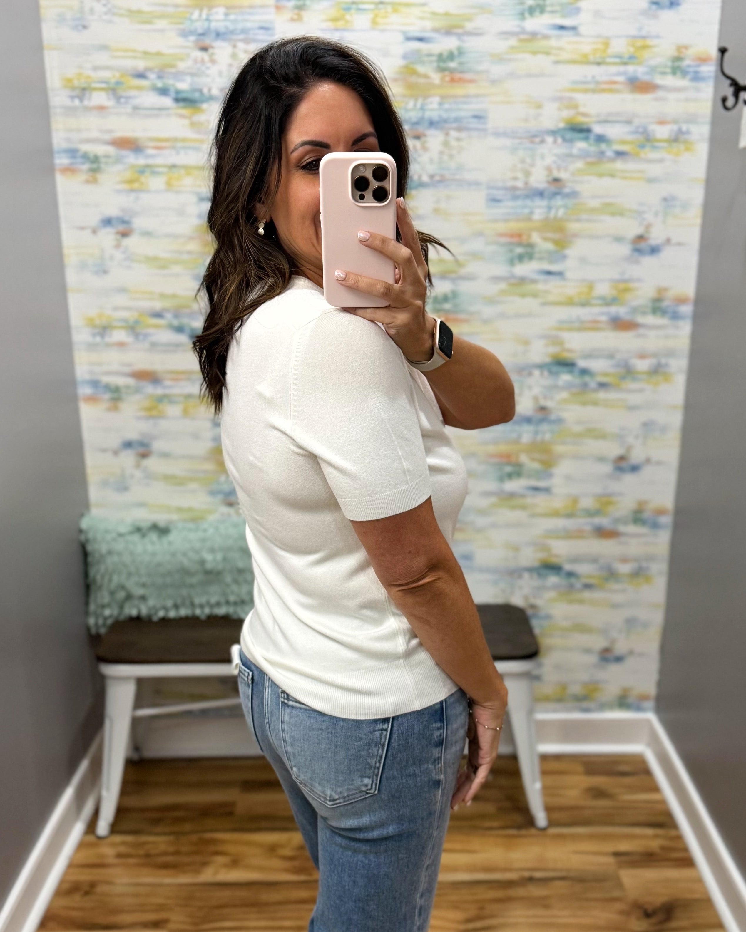 A woman takes a mirror selfie in a fitting room wearing a fitted white short-sleeve top and blue jeans.