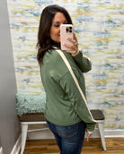 A woman takes a mirror selfie wearing an olive green long-sleeve bomber jacket with cream ribbed cuffs and collar, paired with blue jeans and a cream crossbody bag.