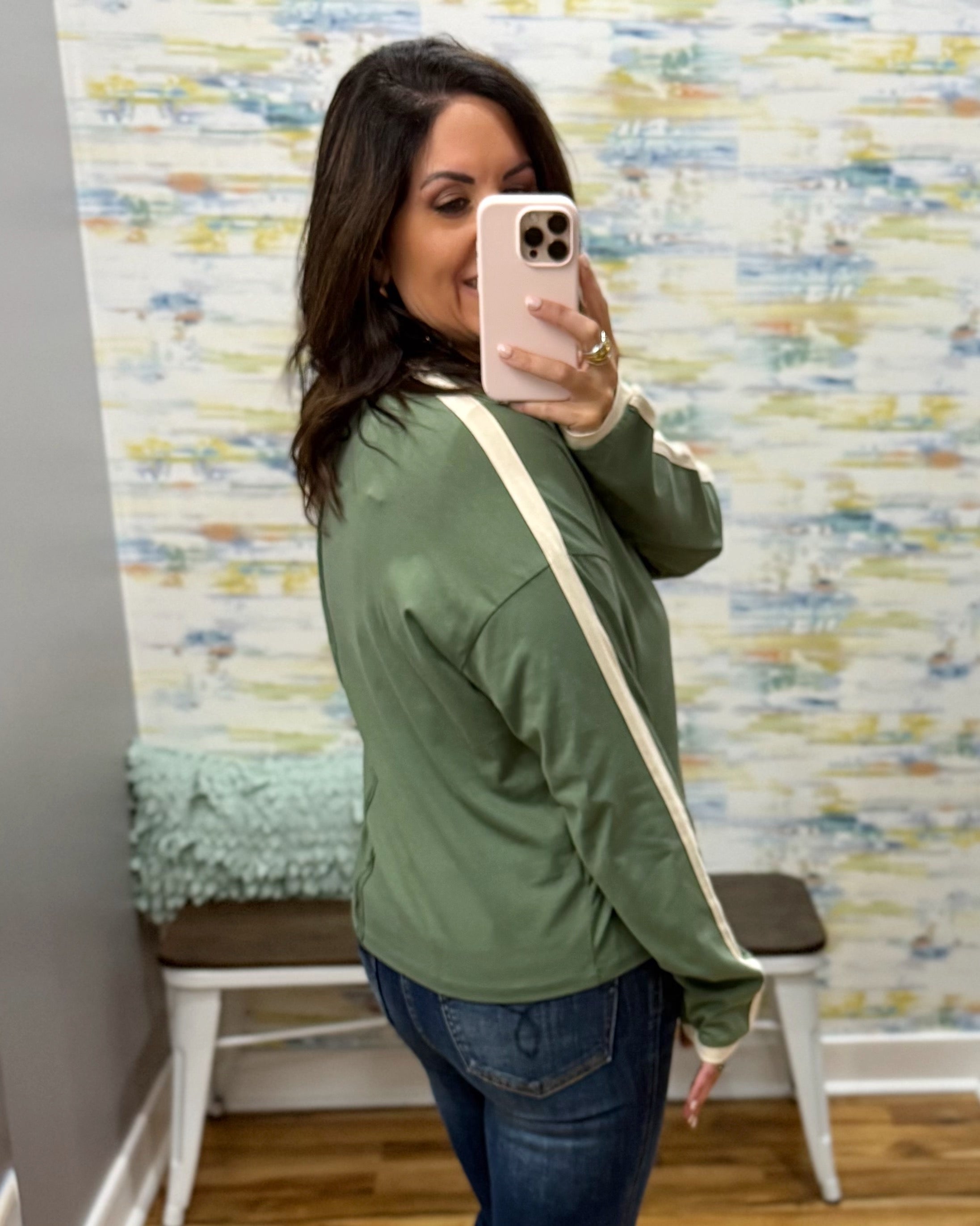 A woman takes a mirror selfie wearing an olive green long-sleeve bomber jacket with cream ribbed cuffs and collar, paired with blue jeans and a cream crossbody bag.