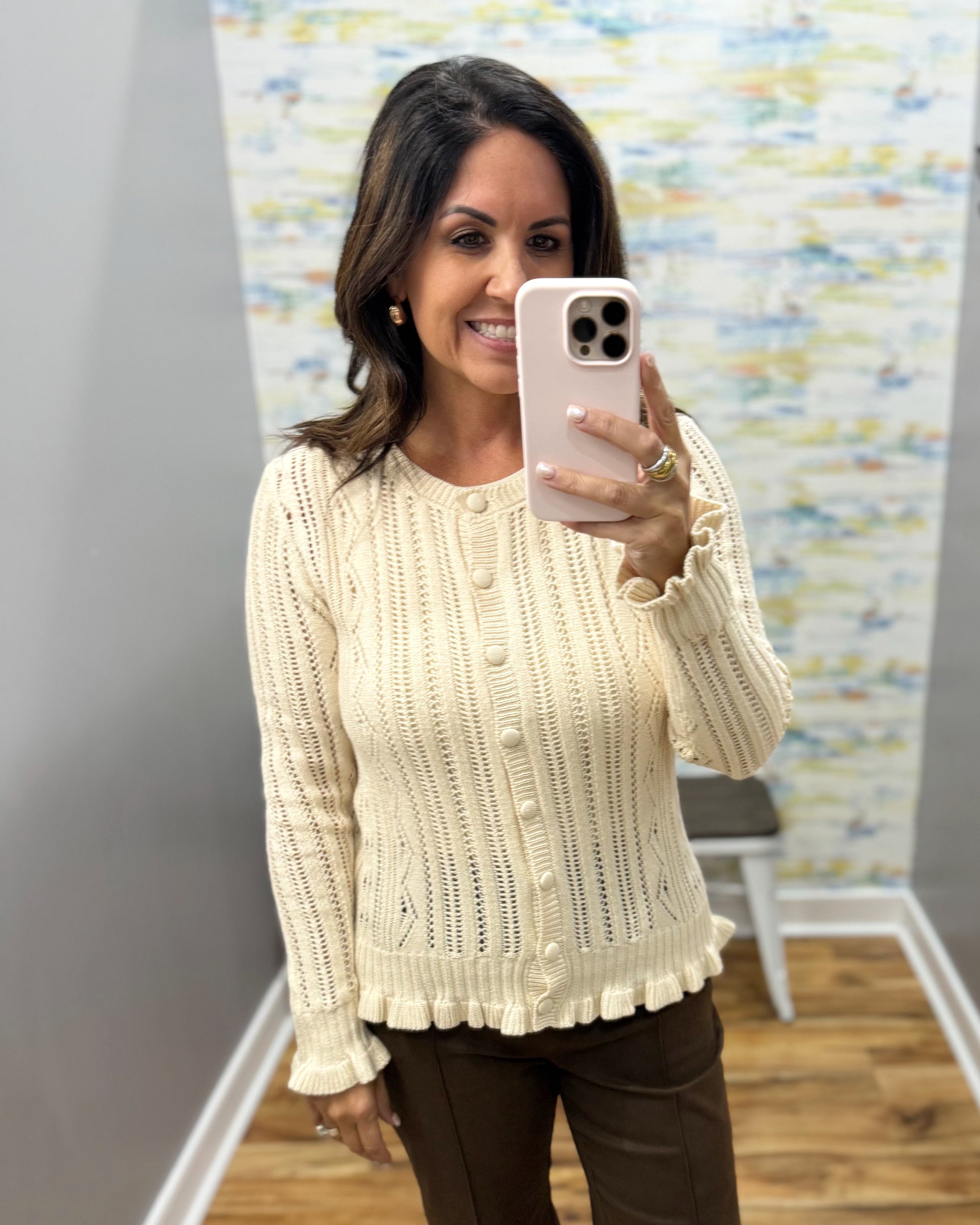 A woman taking a mirror selfie wearing a cream cable-knit button-front cardigan sweater and brown pants in an indoor room with a patterned wallpaper background.