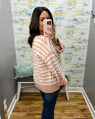 A woman takes a mirror selfie wearing a pink and white striped knit sweater and dark blue jeans in a room with patterned wallpaper and a wooden floor.