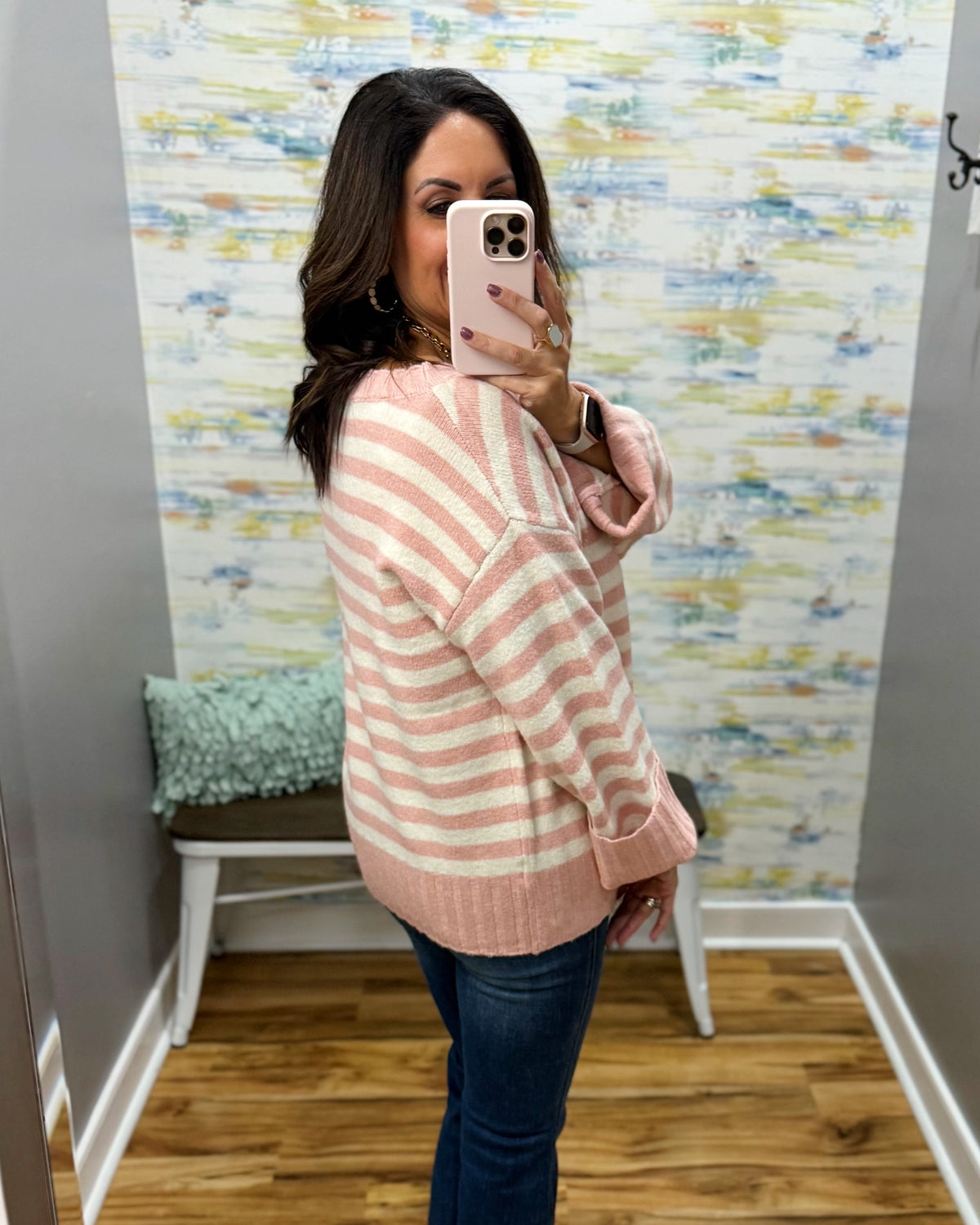 A woman takes a mirror selfie wearing a pink and white striped knit sweater and dark blue jeans in a room with patterned wallpaper and a wooden floor.
