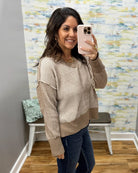 A woman takes a mirror selfie wearing a beige and brown vertically ribbed knit sweater with a crew neck and drop shoulders, paired with dark blue jeans in an indoor room with a patterned wallpaper background.