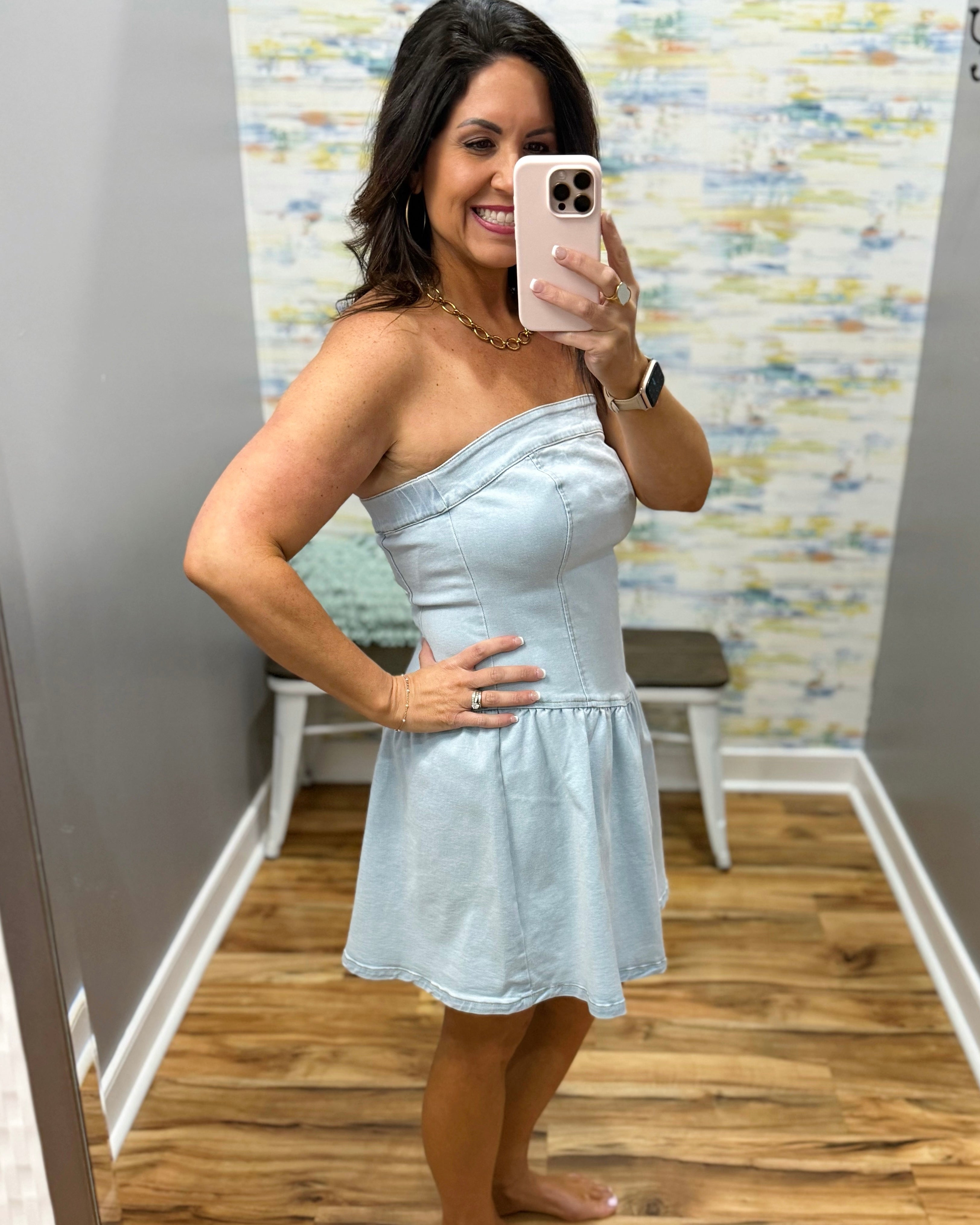Woman taking a mirror selfie wearing a light blue one-shoulder dress in a room with a patterned wall.