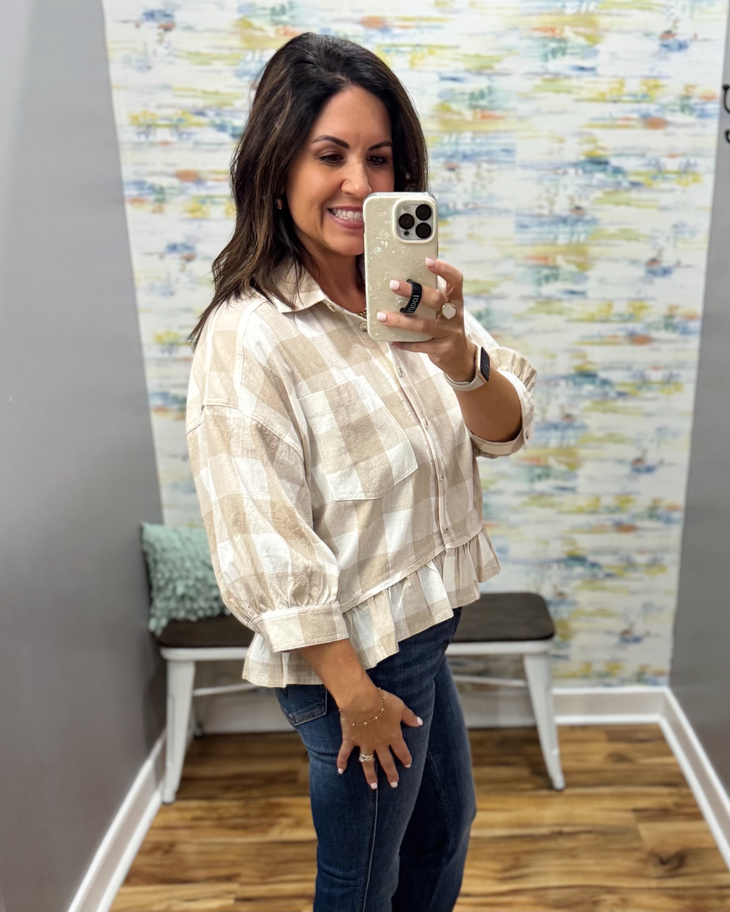 A woman takes a mirror selfie in a fitting room wearing a beige and white plaid button-up shirt with dark blue jeans.