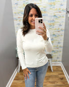 A woman taking a mirror selfie in a fitting room wearing a fitted long-sleeve white top and blue jeans.