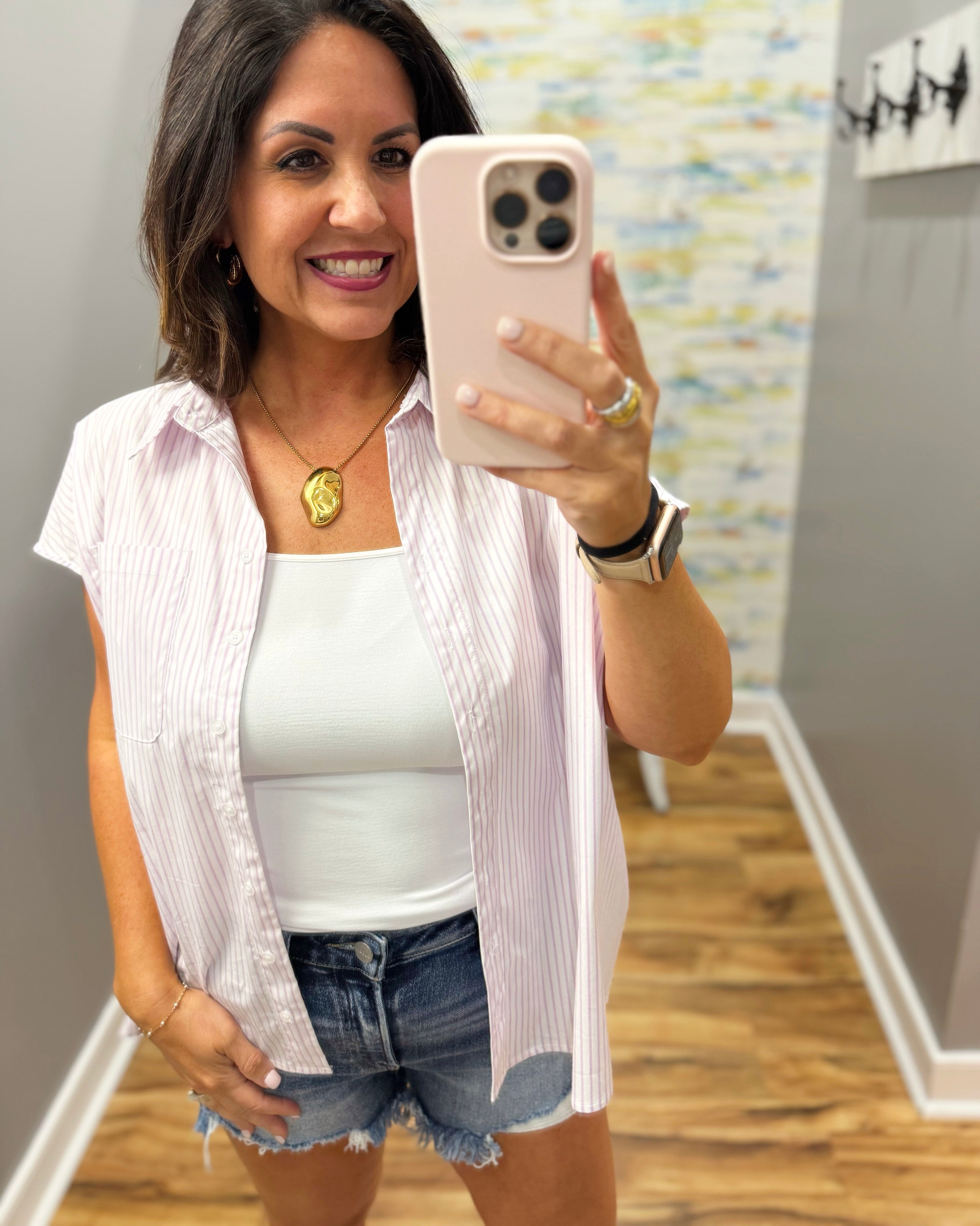 Woman taking a mirror selfie wearing a white top, light pink cardigan, and denim shorts.